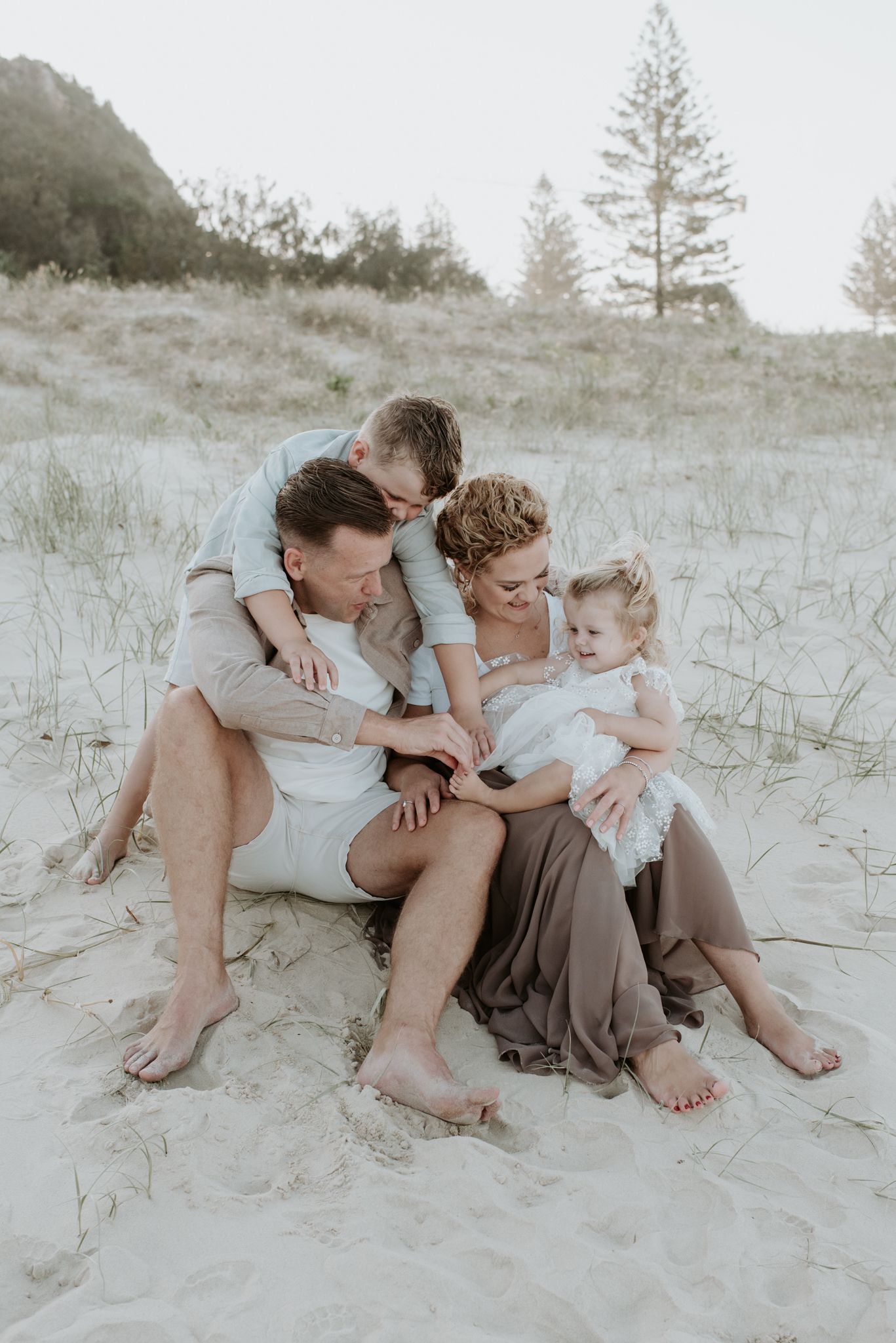 Intimate family moment captured on sandy dunes with pine trees in background.