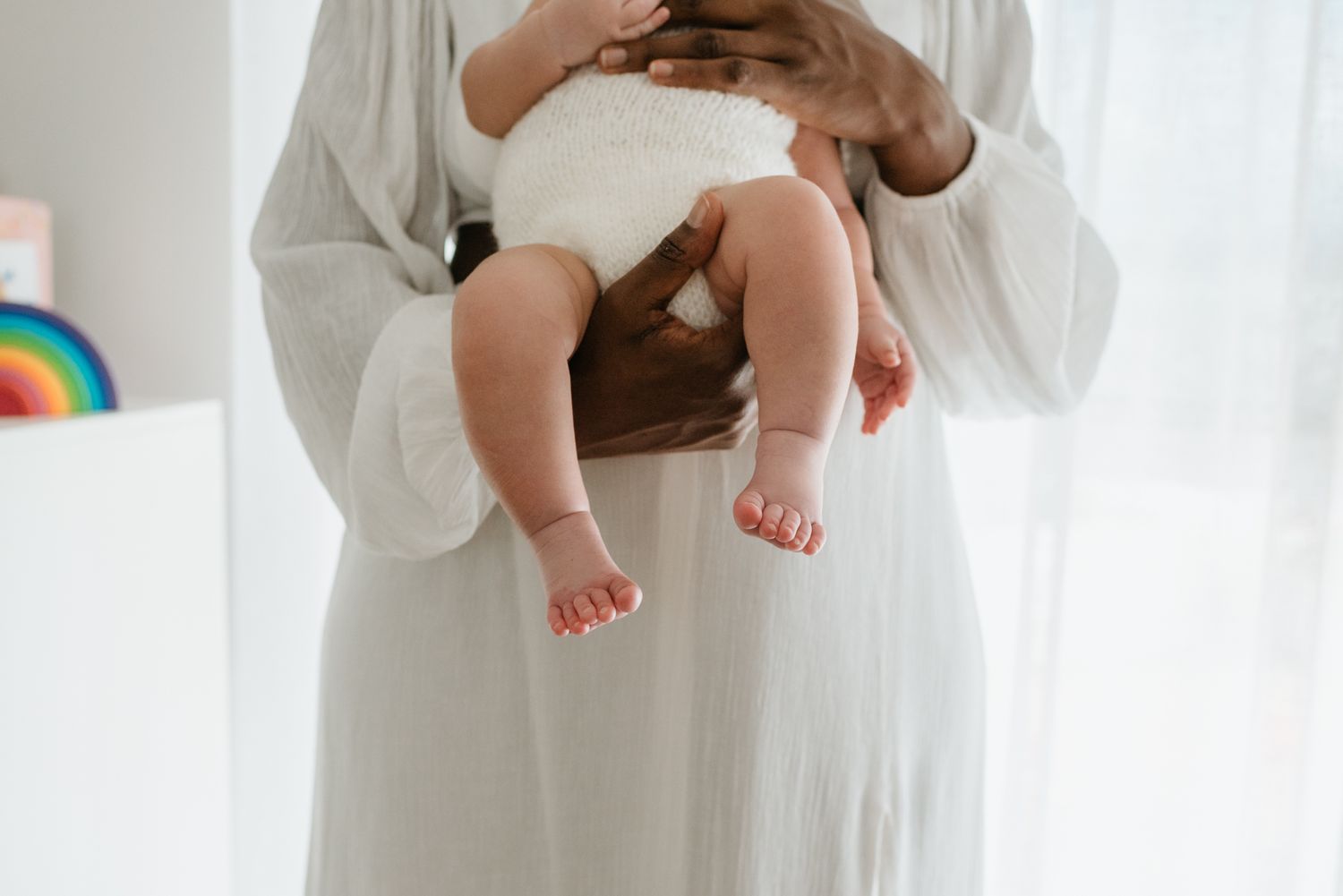A pair of tiny bare baby feet dangle from white fabric while being held by gentle hands at home.