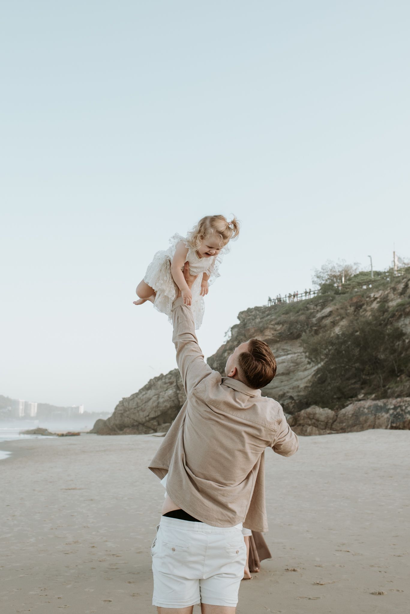 Parent lifting child up high on sandy beach at sunset with coastal cliffs in background.