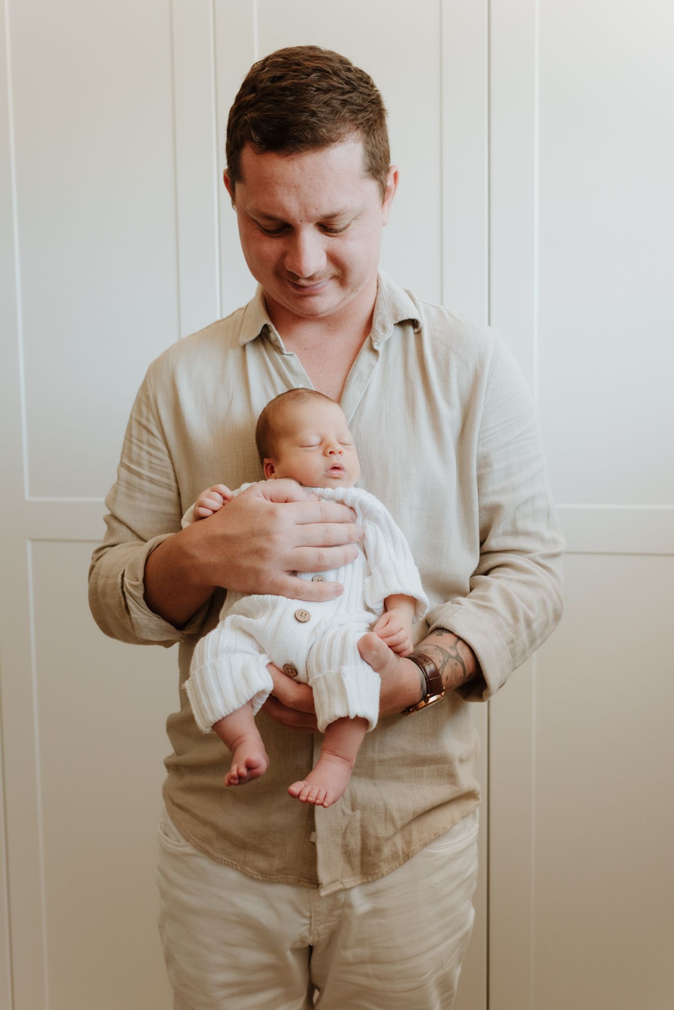 Parent wearing beige shirt holds sleeping newborn baby in white onesie against white wall background.