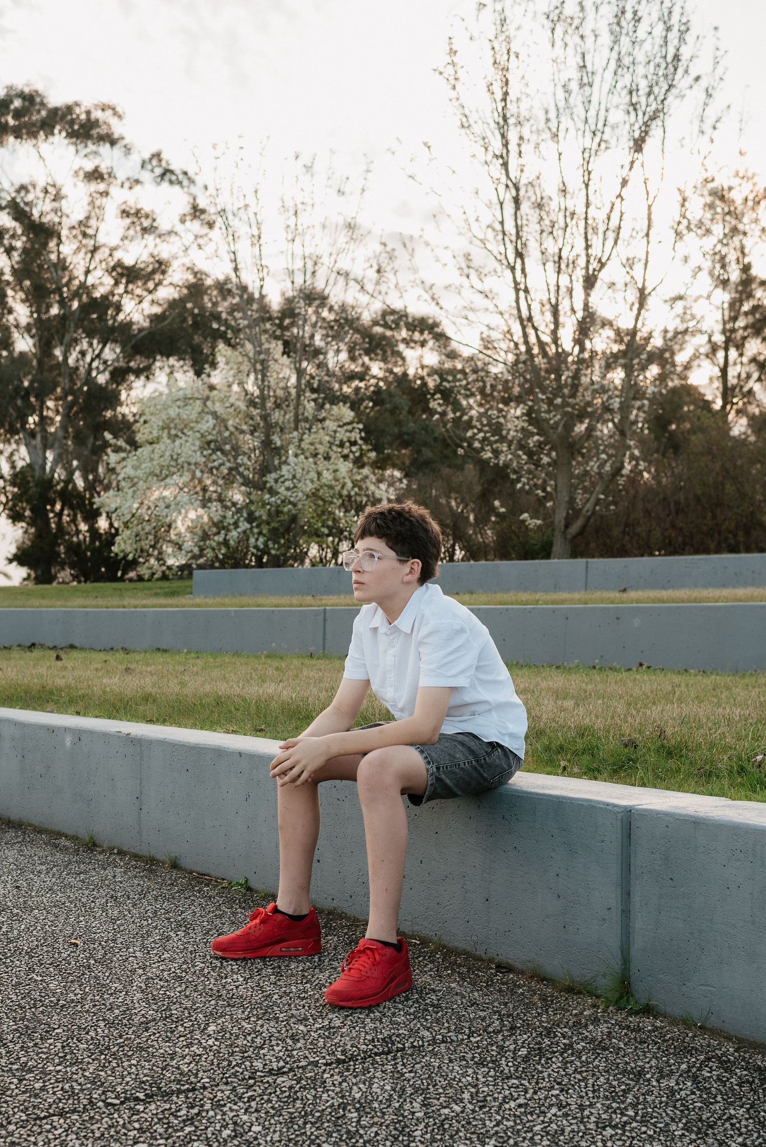 A young boy in a white shirt, gray shorts, and red sneakers sits on a concrete ledge outdoors, gazing thoughtfully.