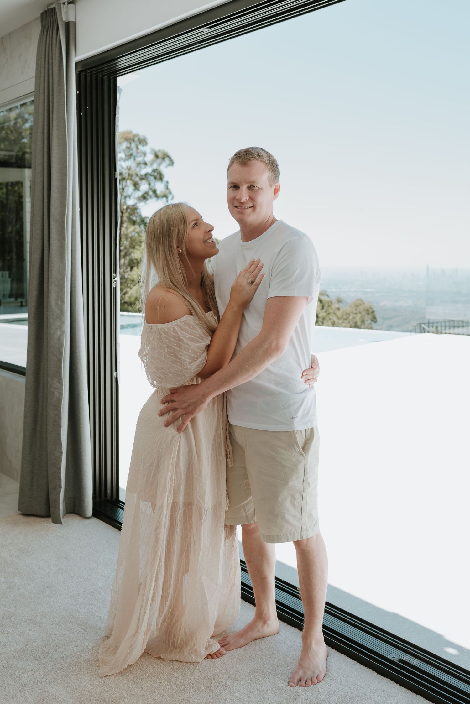 Couple embracing by floor-to-ceiling windows with city views, woman in flowing dress, man in white tee and shorts.