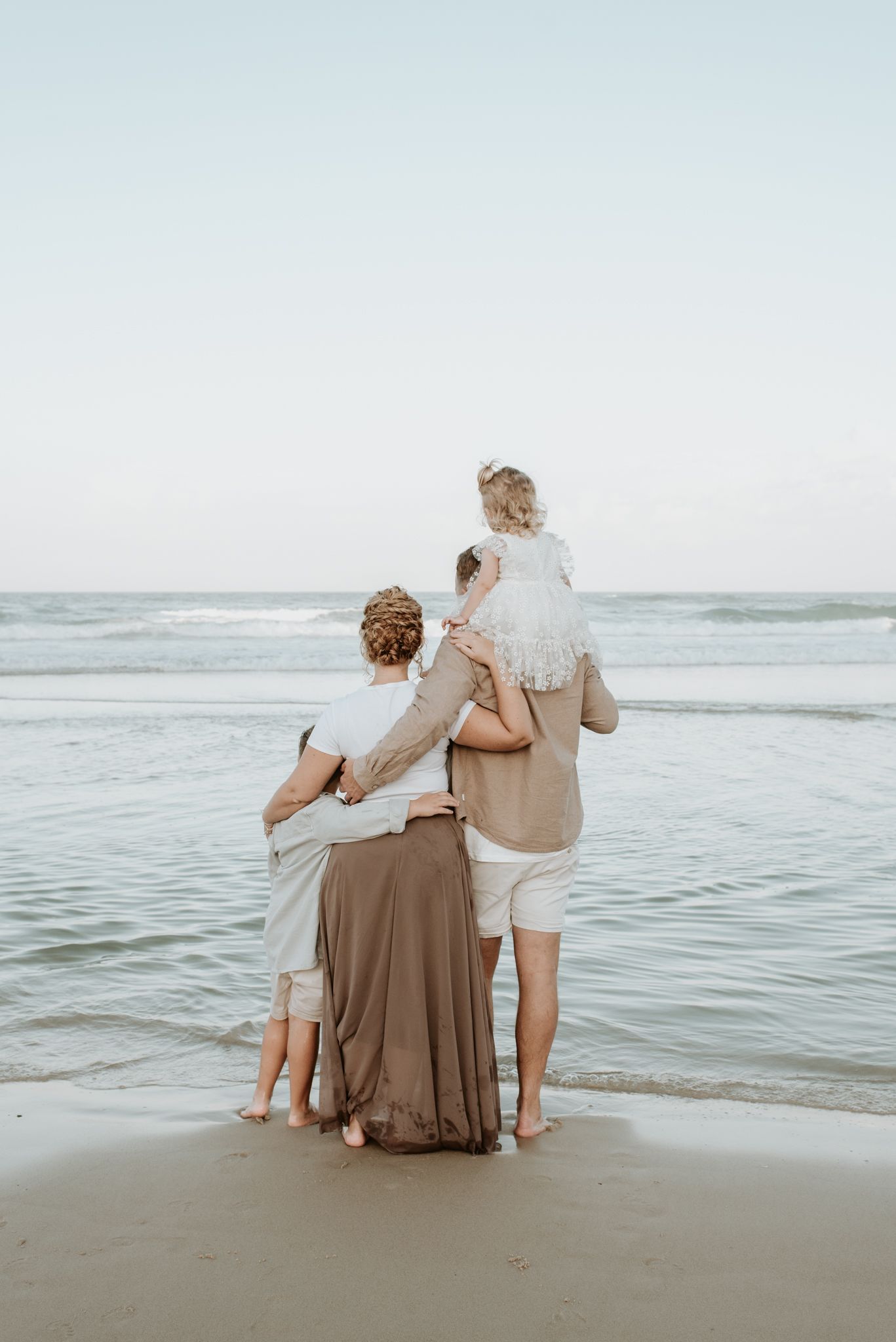 A family stands together at the edge of the ocean, wearing neutral colored clothing while looking out at the water.