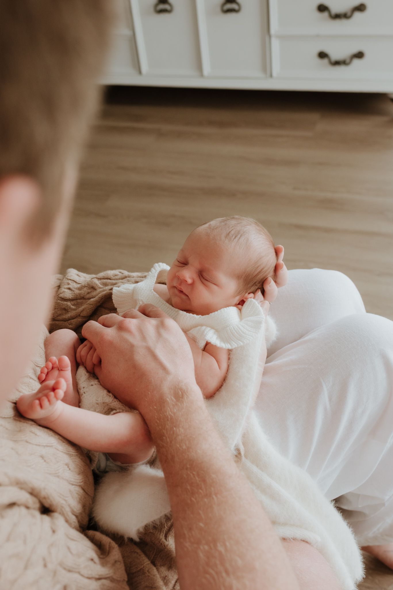 A father tenderly holds his sleeping newborn baby dressed in white on a cozy blanket on the floor.