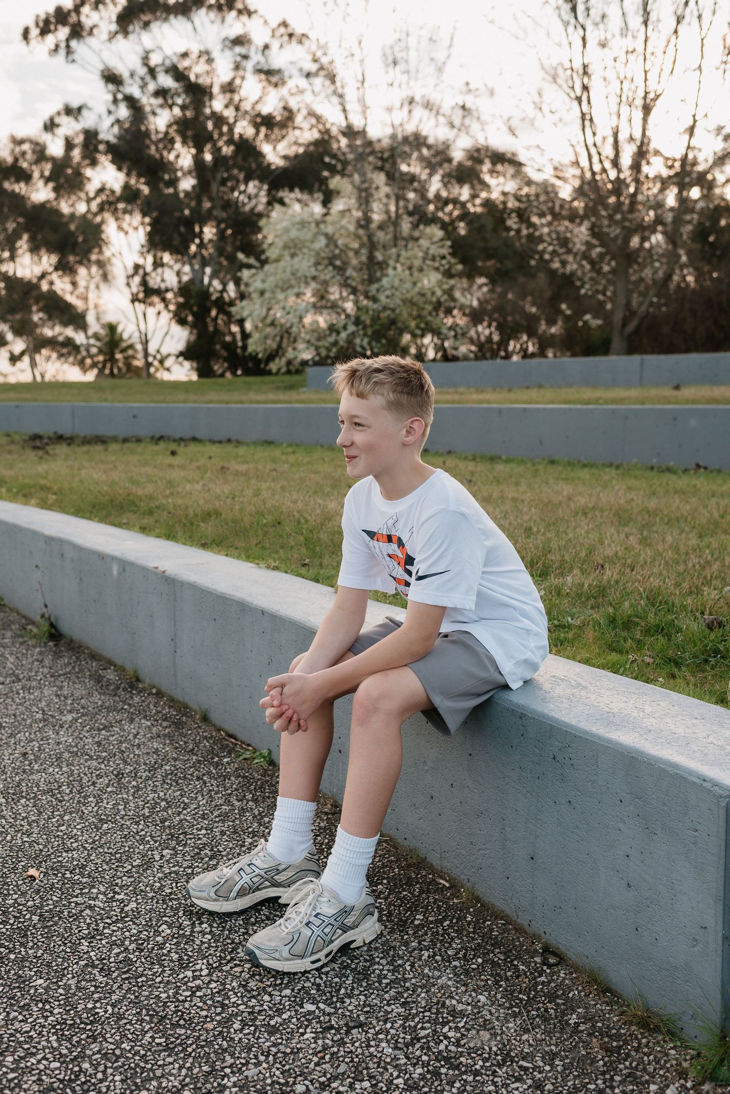 A smiling boy sits on a concrete ledge outdoors, wearing a white t-shirt, grey shorts, and sneakers.