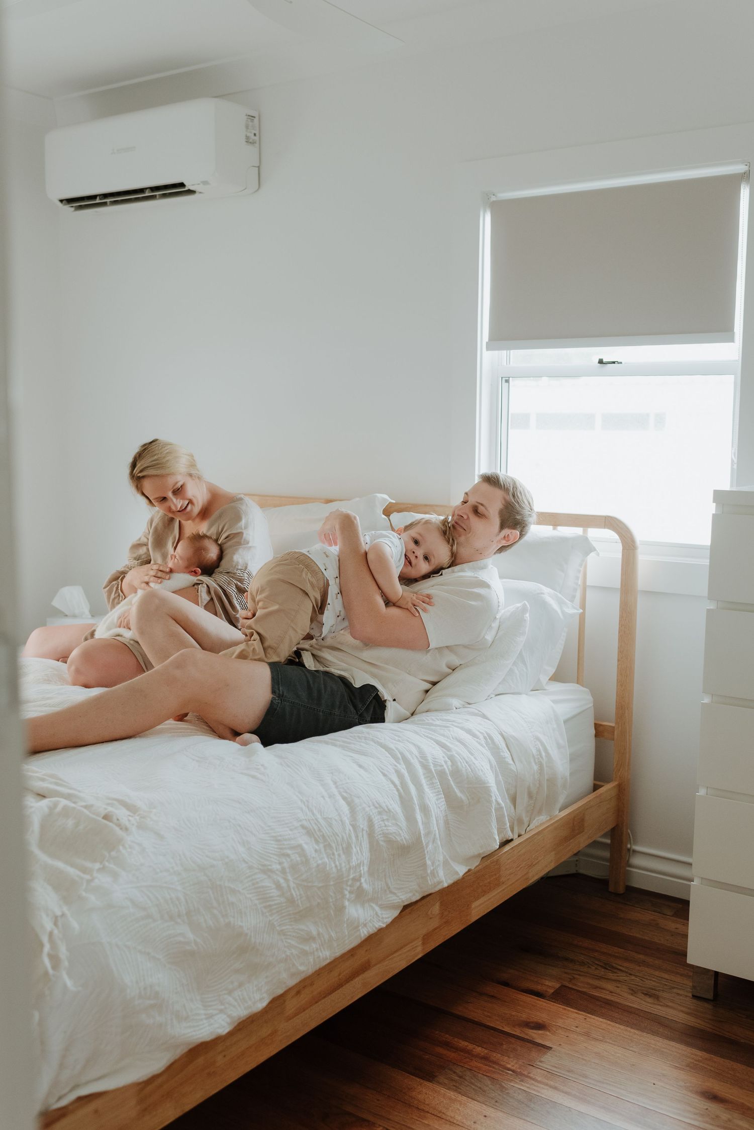 Family relaxes together on wooden bed with white bedding in minimalist bedroom with air conditioning unit.