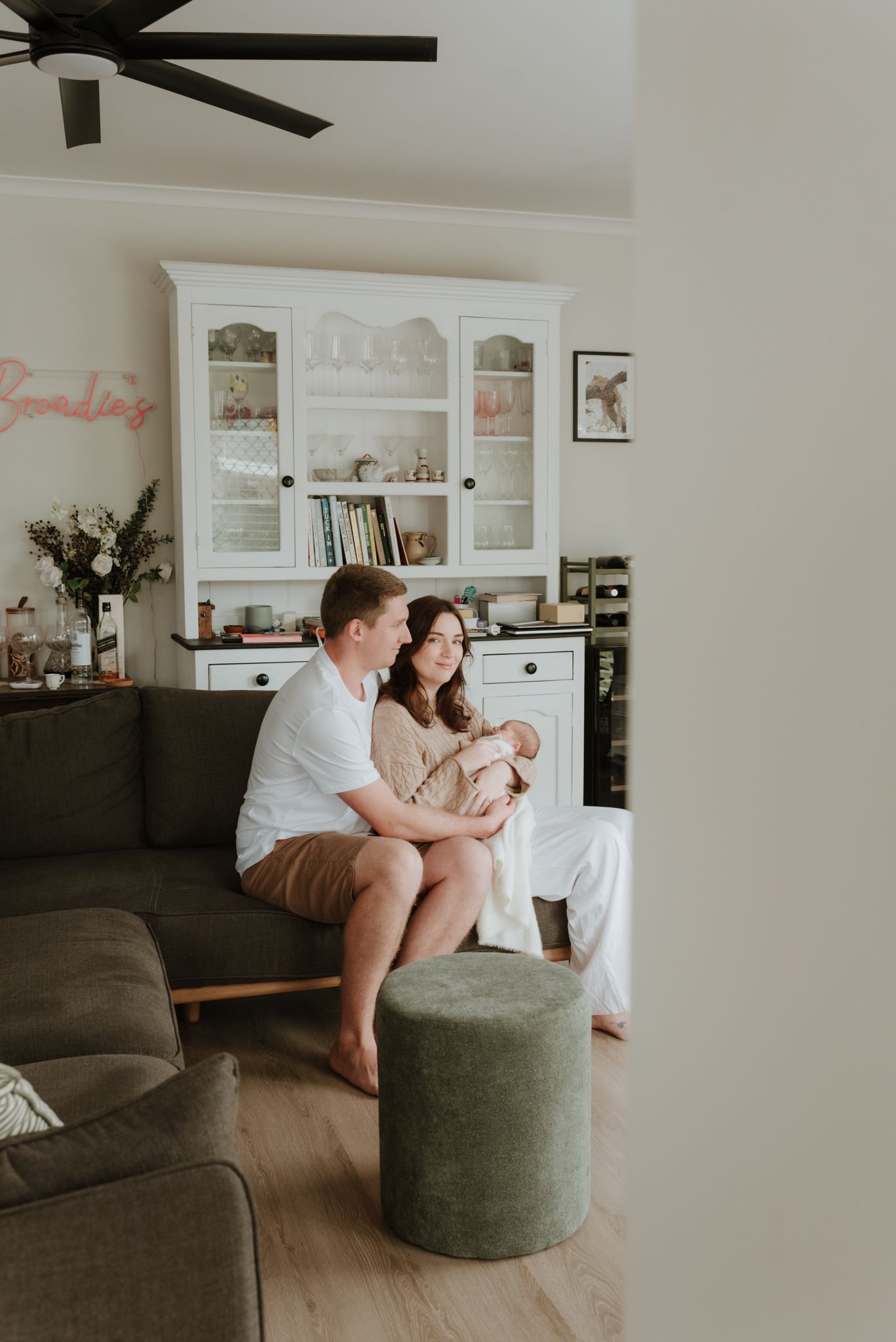 A couple sits on a gray sofa holding their newborn baby in a cozy living room with a white hutch.