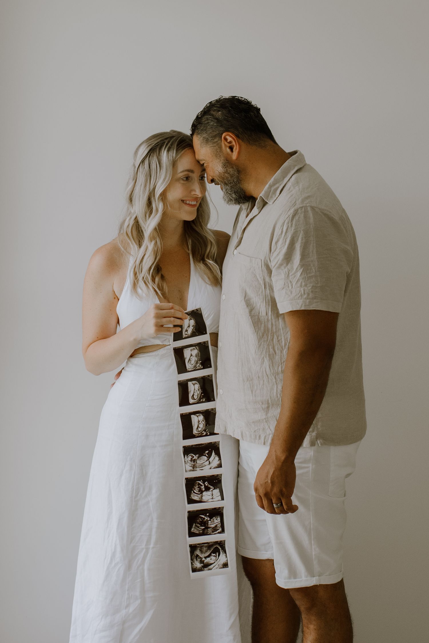 A couple in white clothing shares an intimate moment while holding ultrasound photos in a minimalist setting.