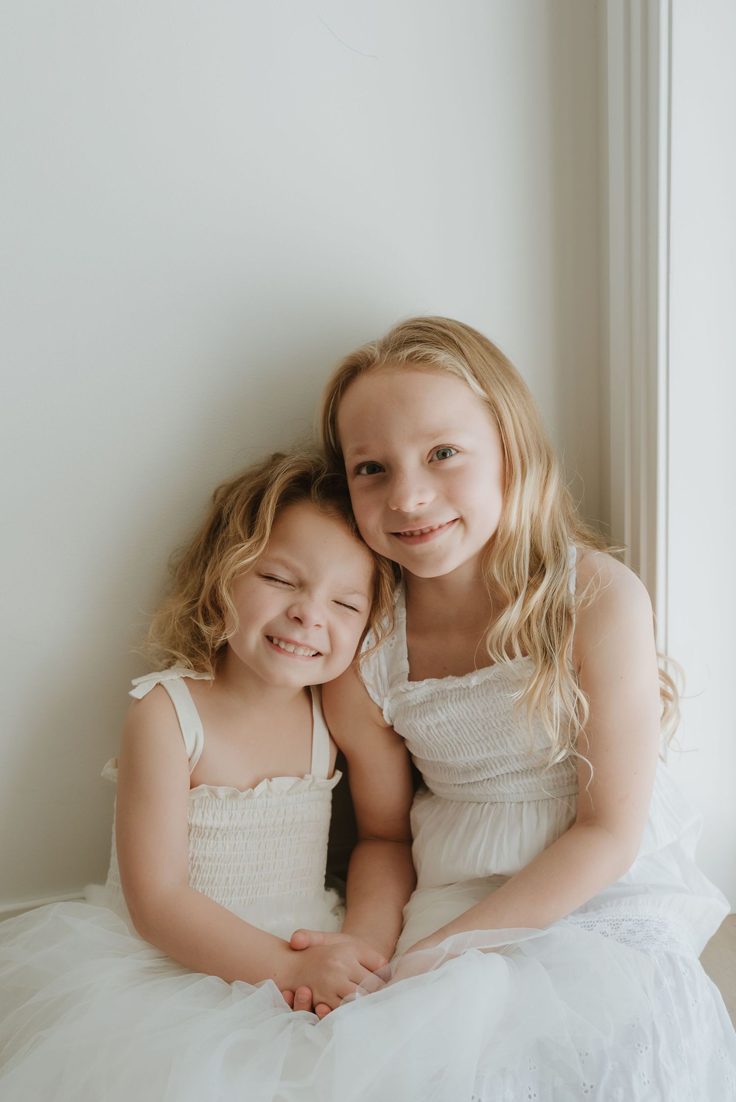 Two young sisters in white dresses smiling and hugging together against a light background.