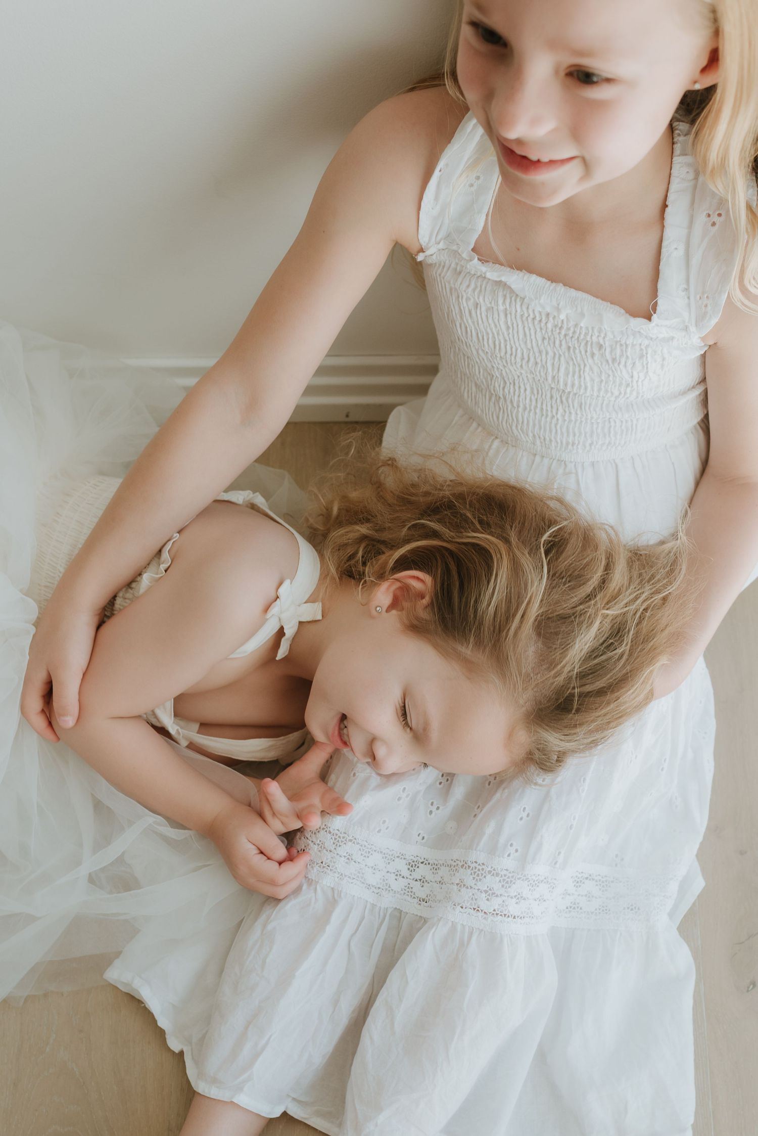 Two young girls in white dresses share a tender moment, with older sister gently embracing younger child.
