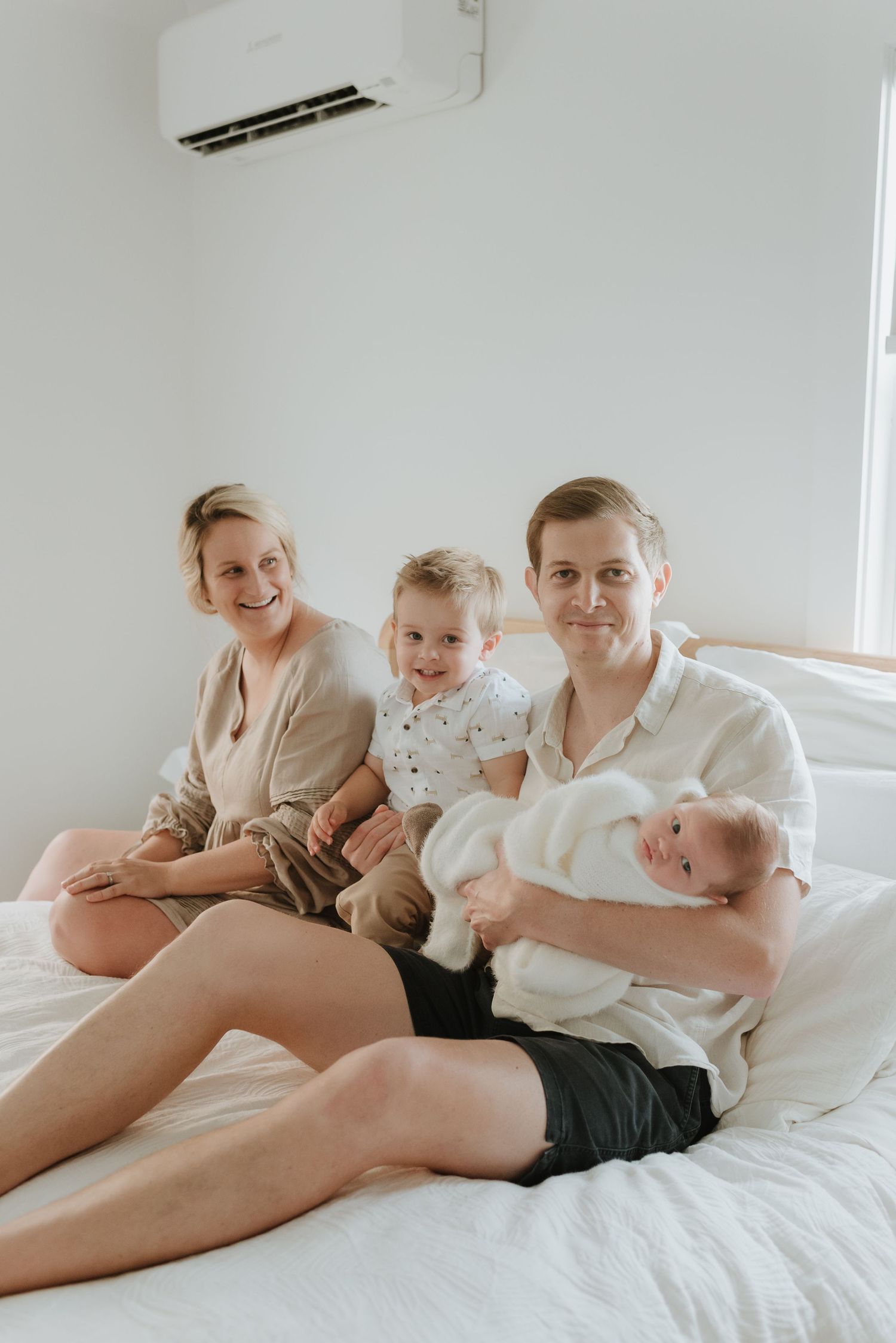 A family relaxes together on a white bed under a wall-mounted air conditioning unit.