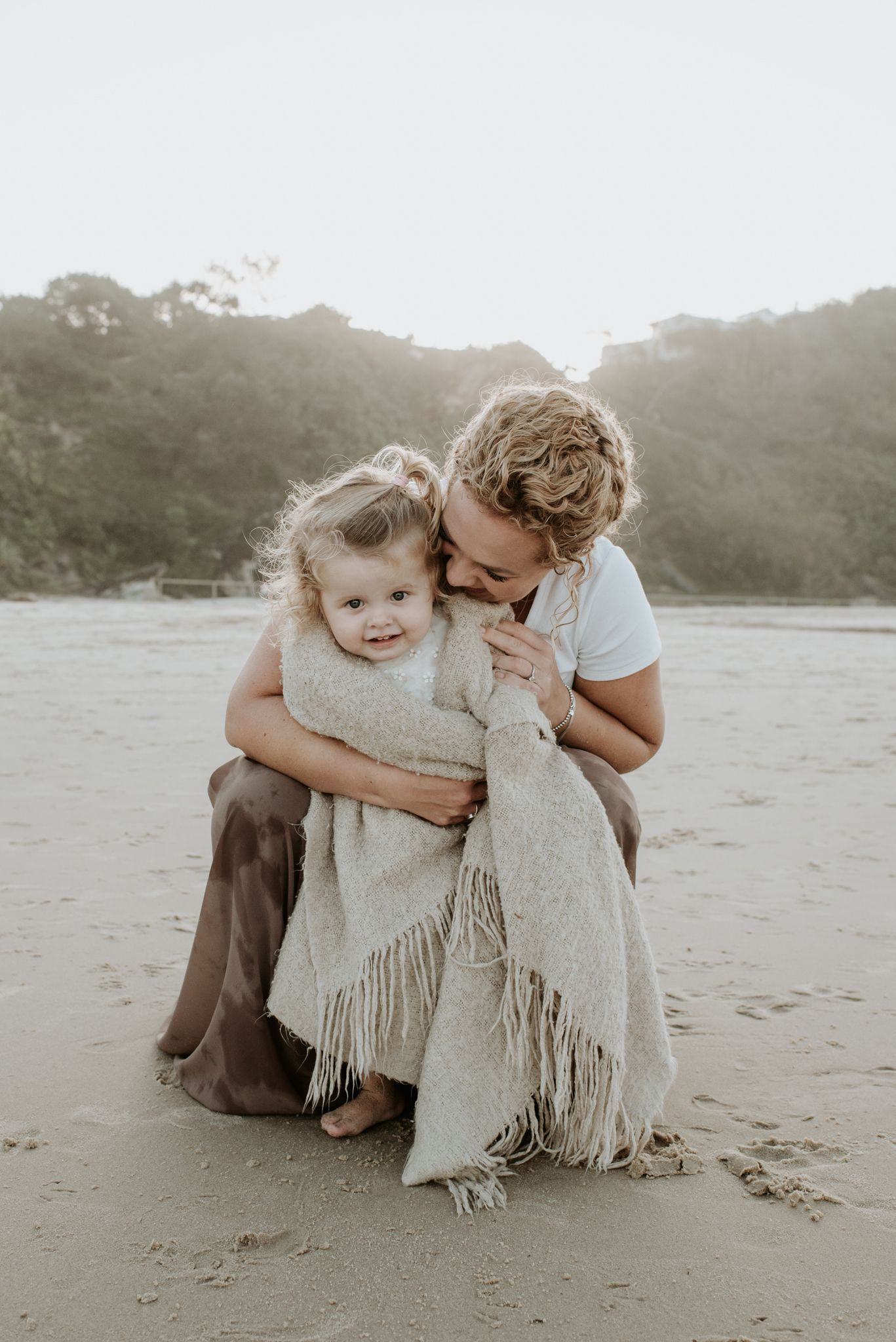 An intimate family moment is shared between parent and child wrapped in a cozy blanket on the beach.
