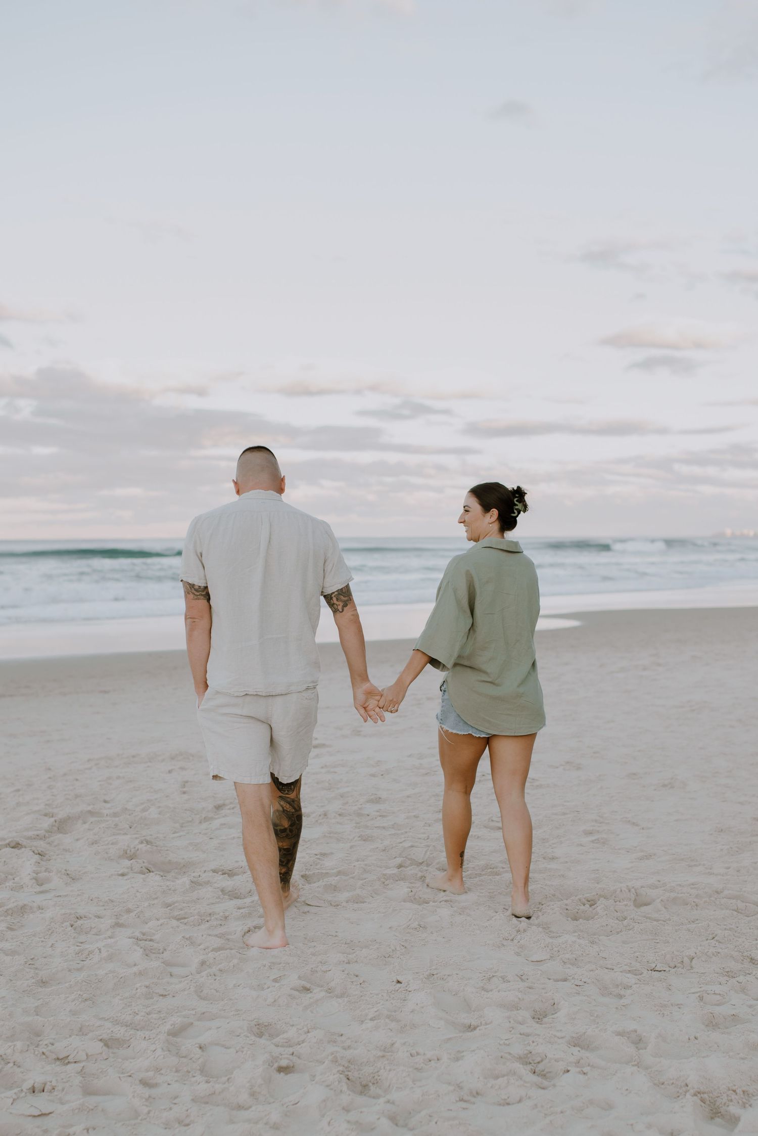 A couple walks hand in hand along a serene beach at dusk with gentle waves and cloudy skies in the background.