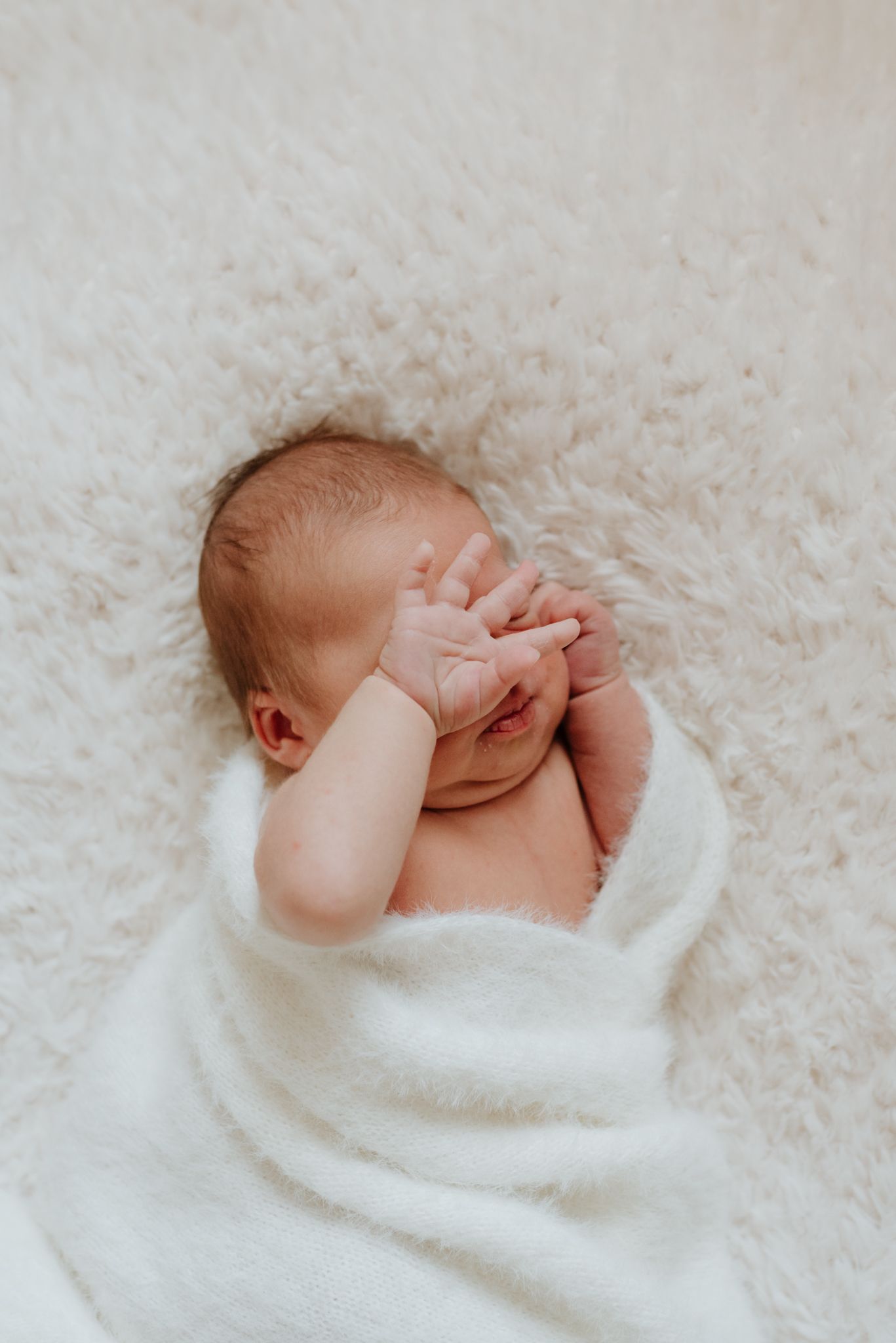 Peaceful newborn baby wrapped in soft white blanket sleeping on fluffy white surface with hand near face.