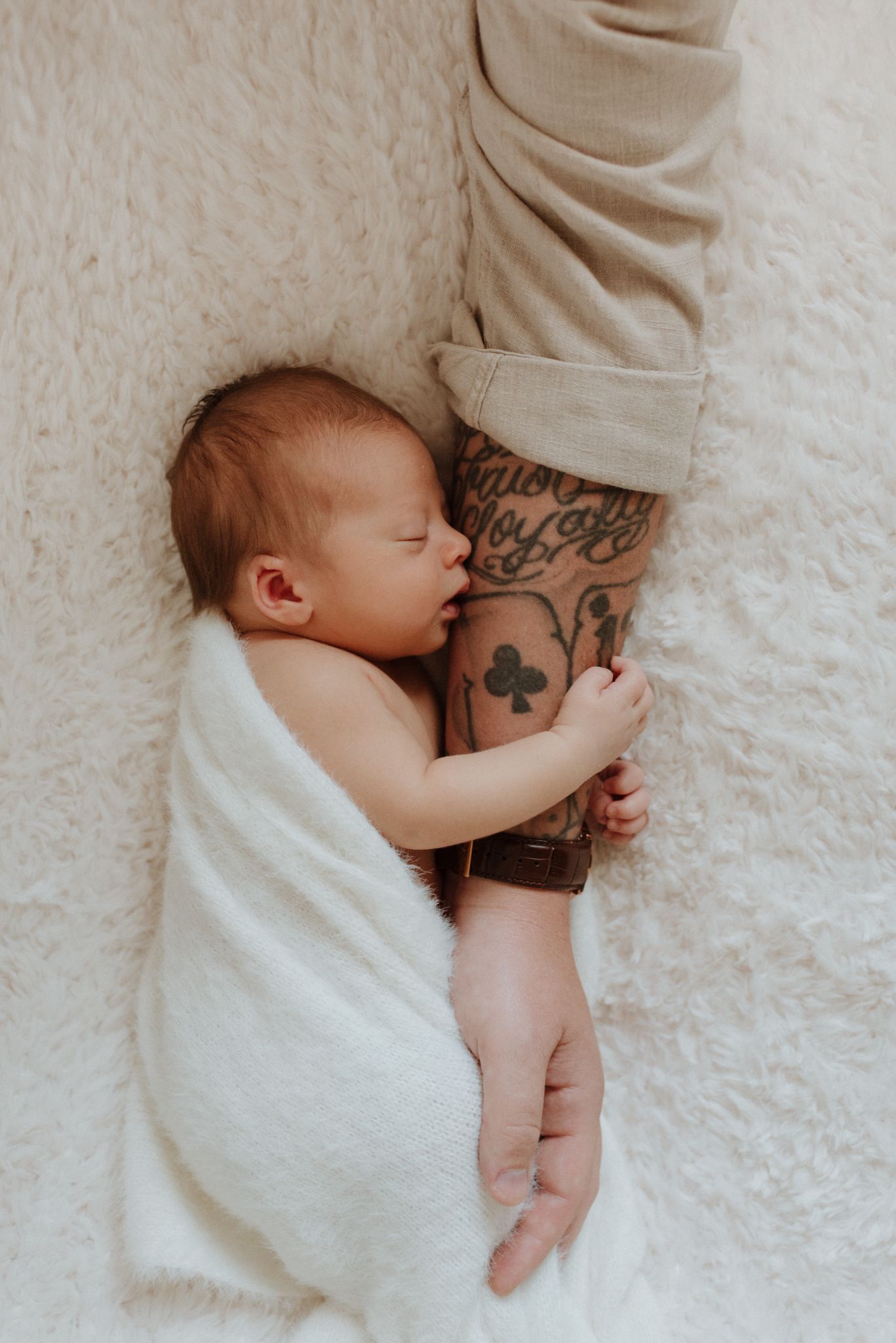 Sleeping baby wrapped in white blanket holding tattooed arm with clover design on white textured background.