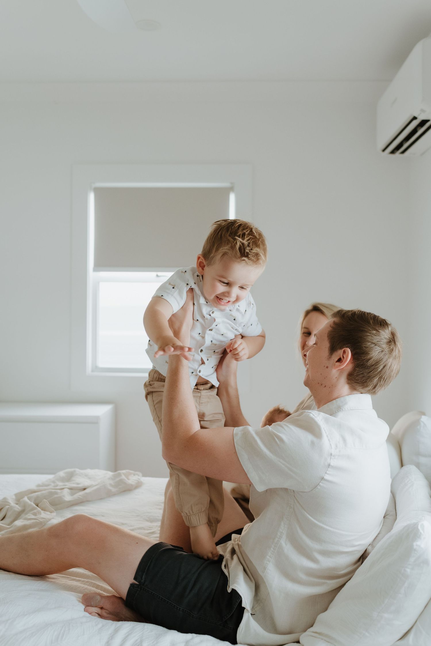 A parent in white shirt plays and lifts up a smiling toddler on a white bed in a bright minimalist room.