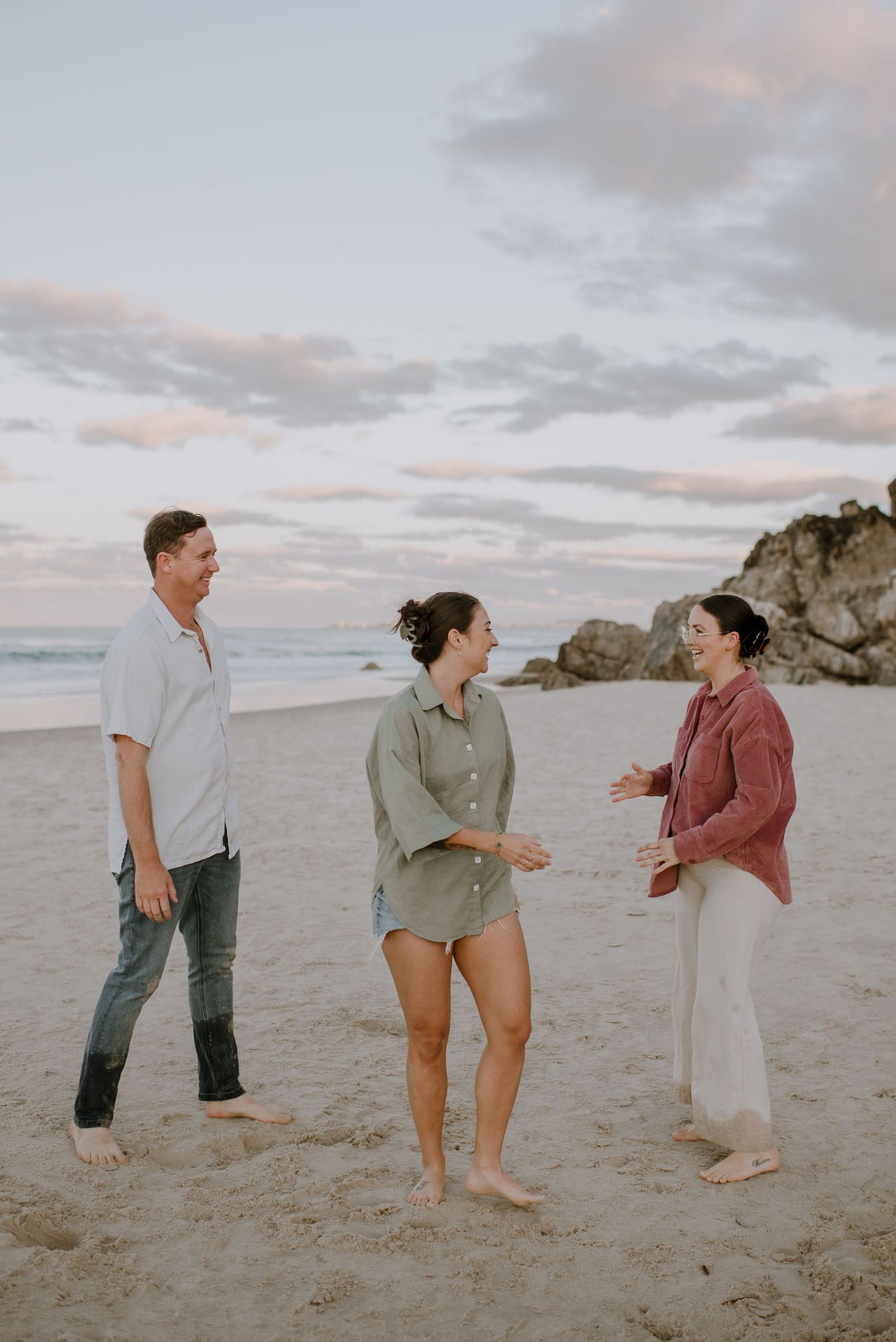 Three people having a casual conversation on a beach at sunset with rocky cliffs in the background.