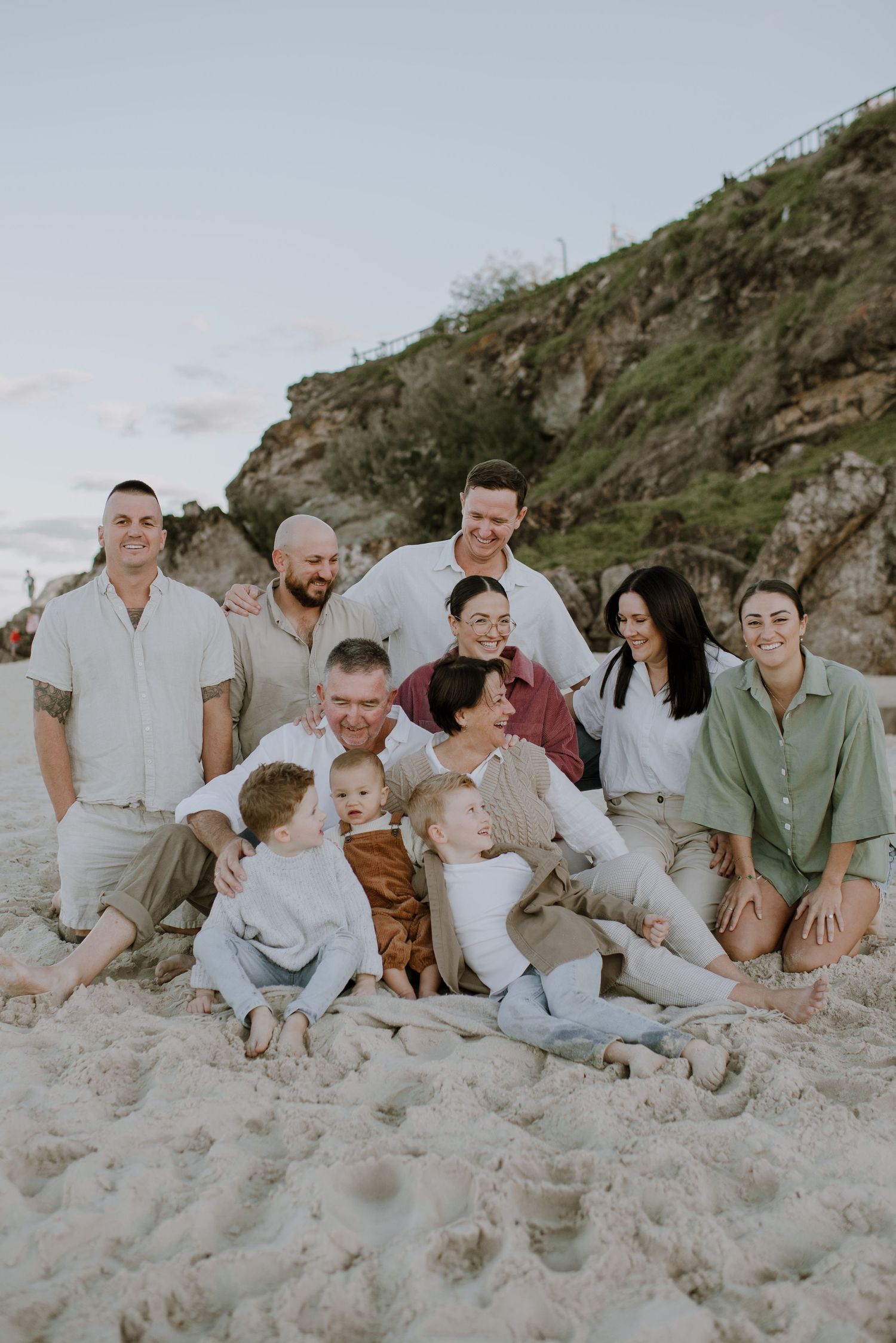A family group wearing neutral colors sits together on a sandy beach with a rocky cliff backdrop at sunset.