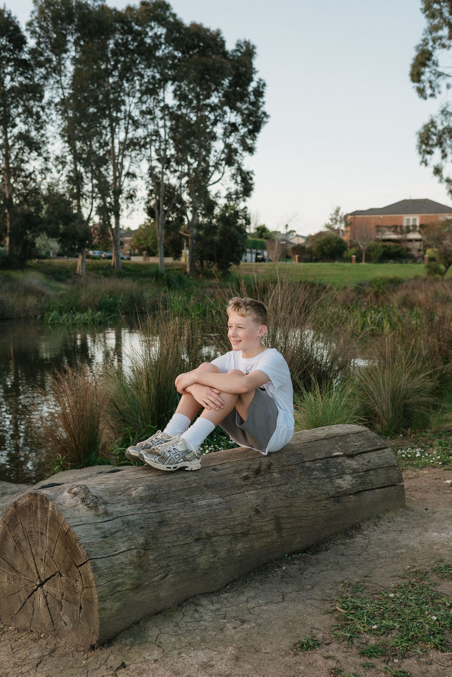 A smiling boy sits on a large log by a peaceful pond, surrounded by lush greenery and trees in a park setting.