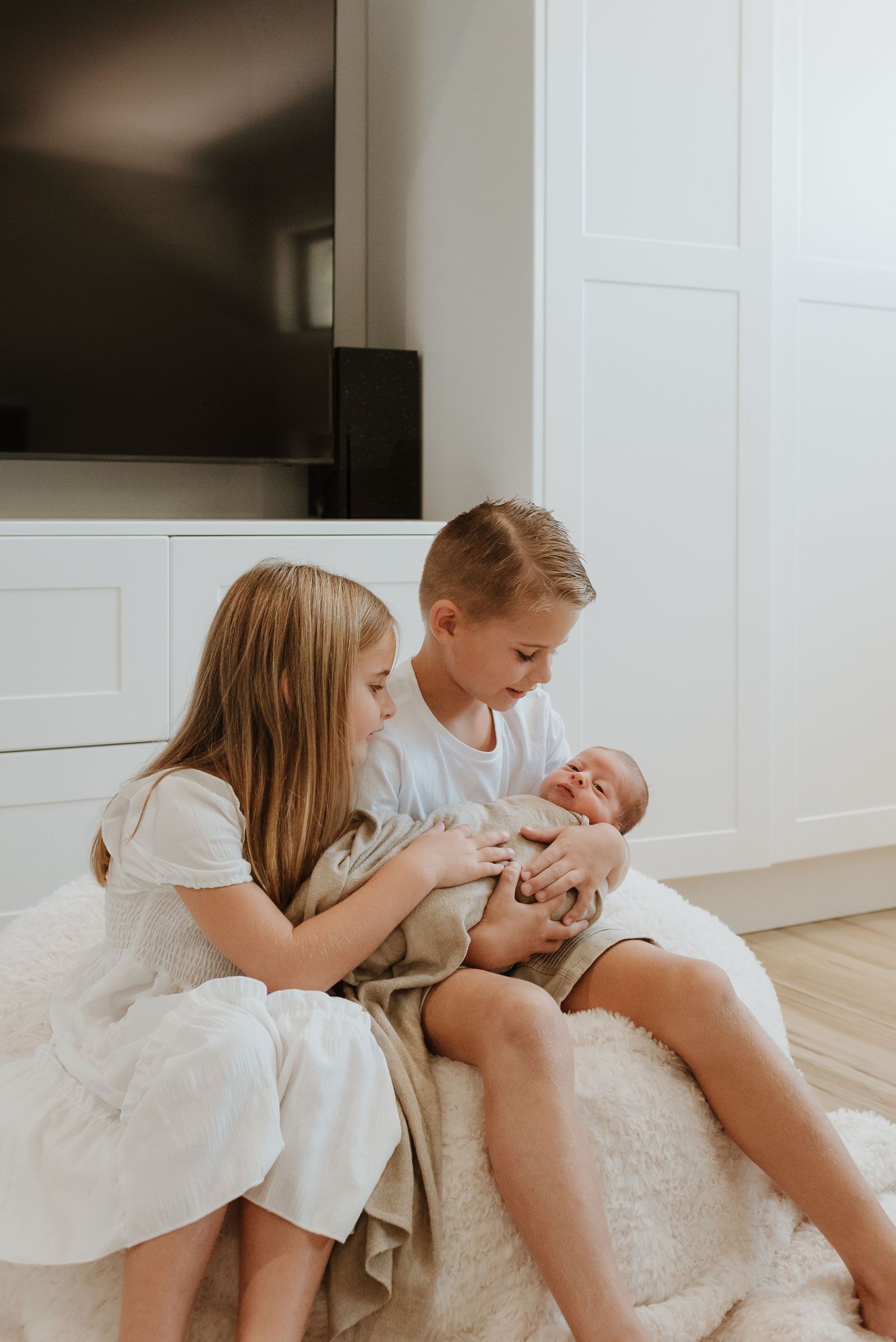 Two young children in white outfits sit on a plush carpet while tenderly looking at and cradling a newborn baby.