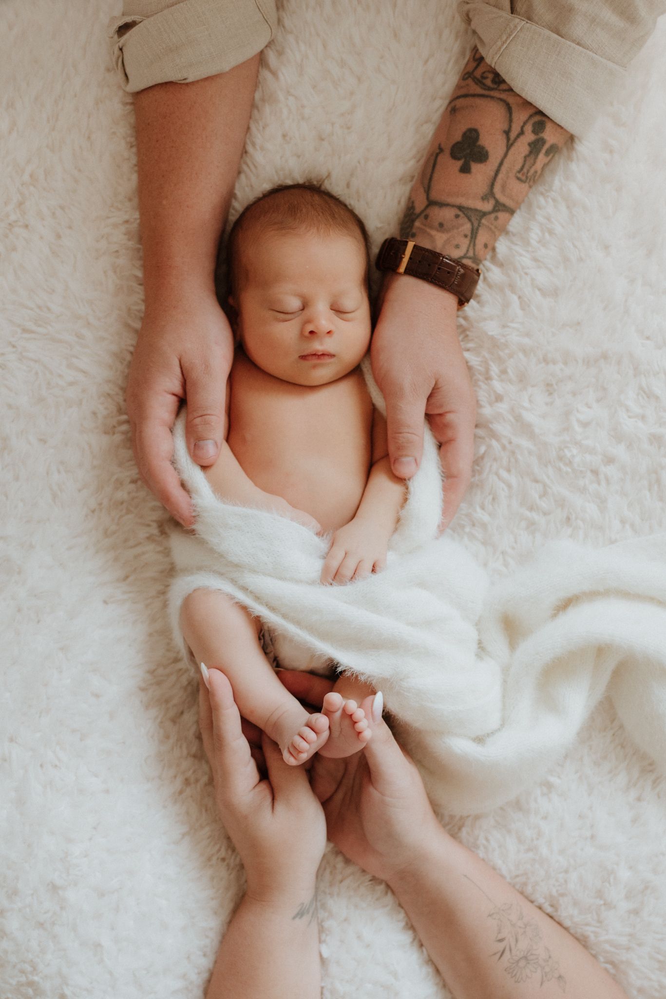 A sleeping newborn baby wrapped in white fabric lies on a blanket while hands gently touch their feet and body.