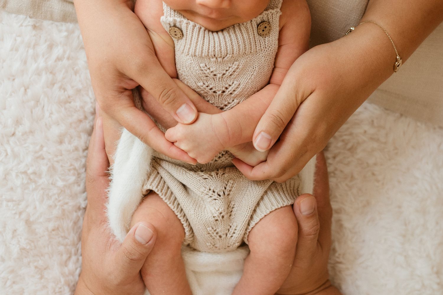 Small feet and legs in a cream knitted romper are gently held by caring hands against a soft textured background.
