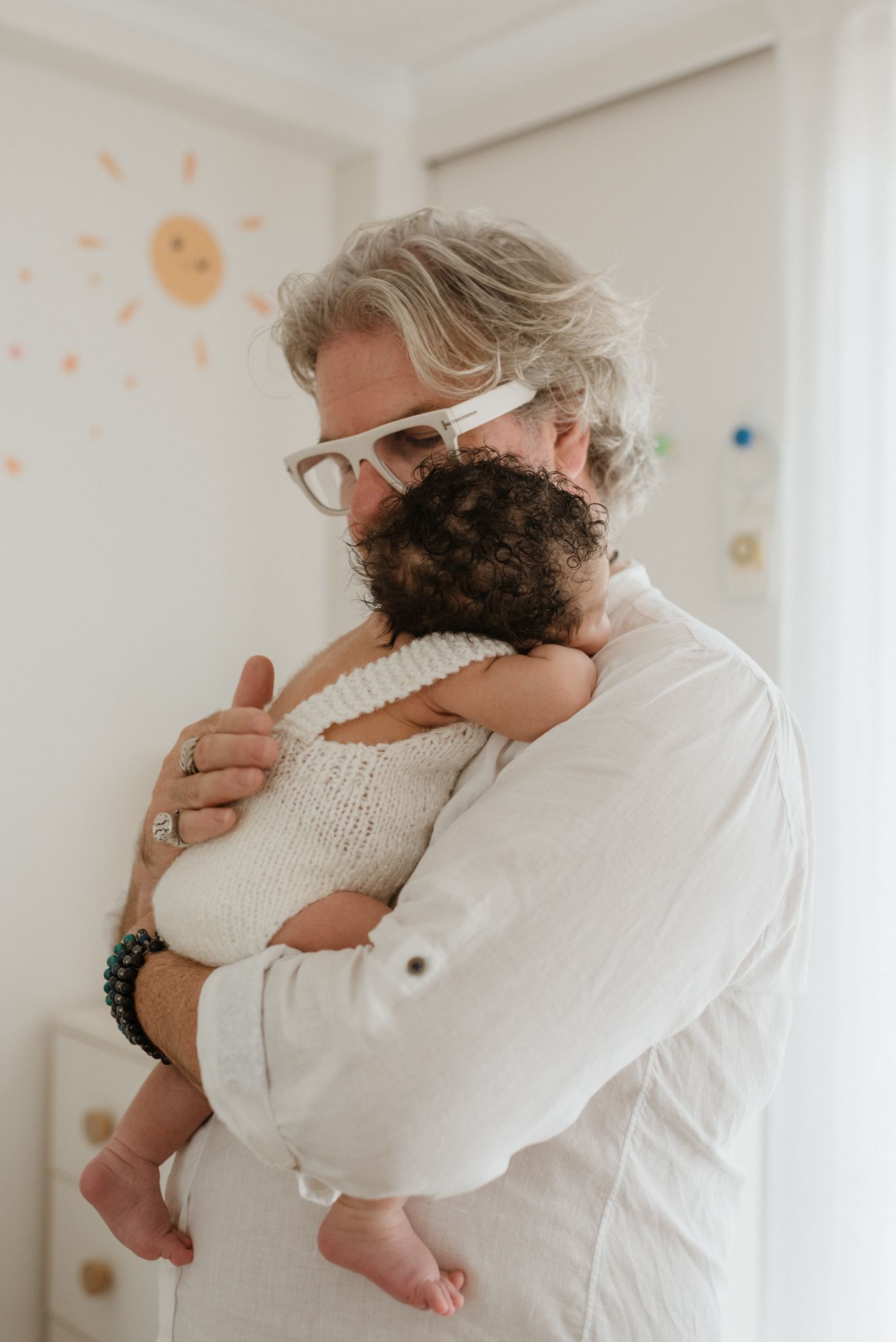 A tender moment between a caregiver in white attire and a baby wearing a knitted outfit, standing against a pastel wall.