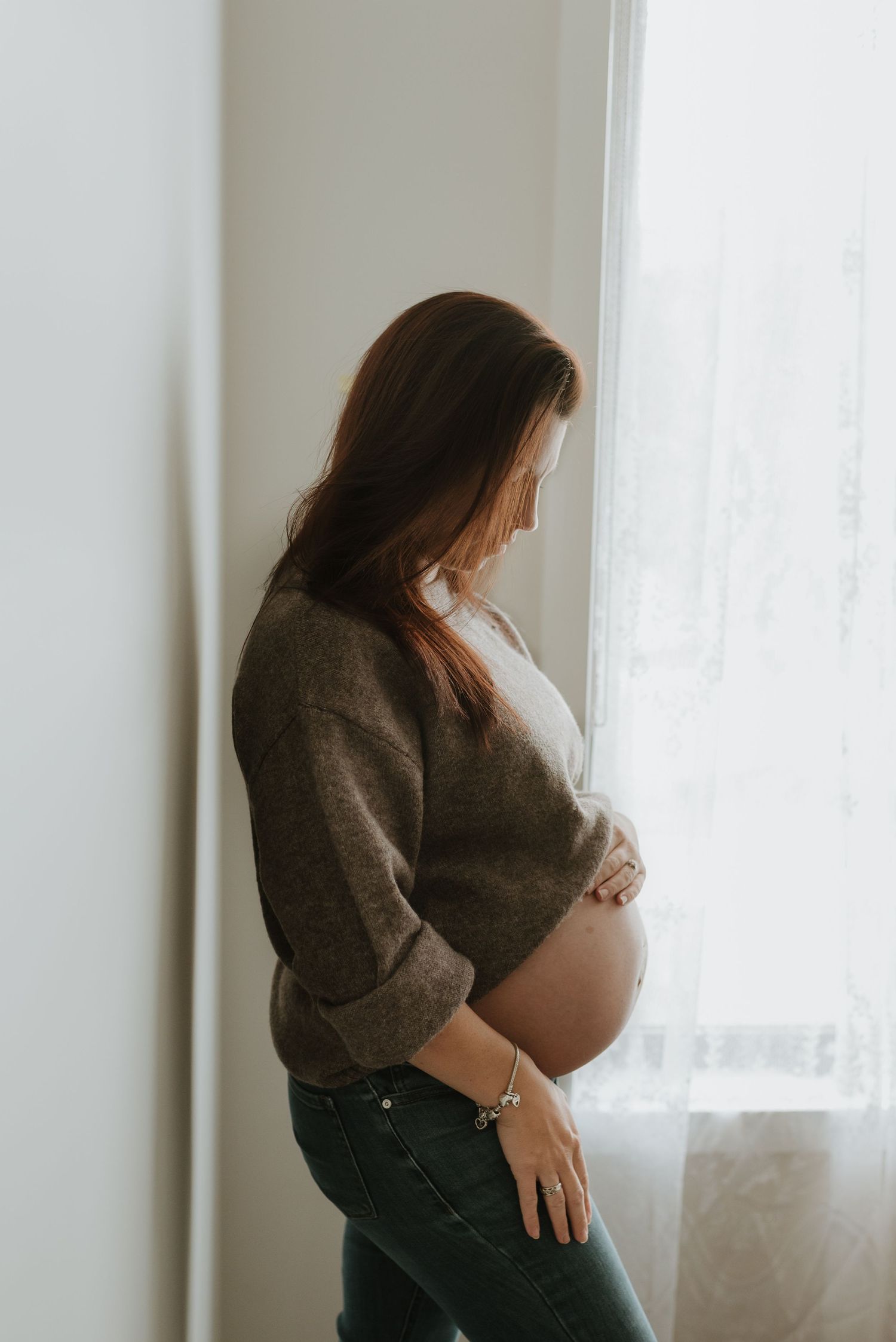 Pregnant woman in gray sweater and jeans cradling her baby bump by a bright window indoors.