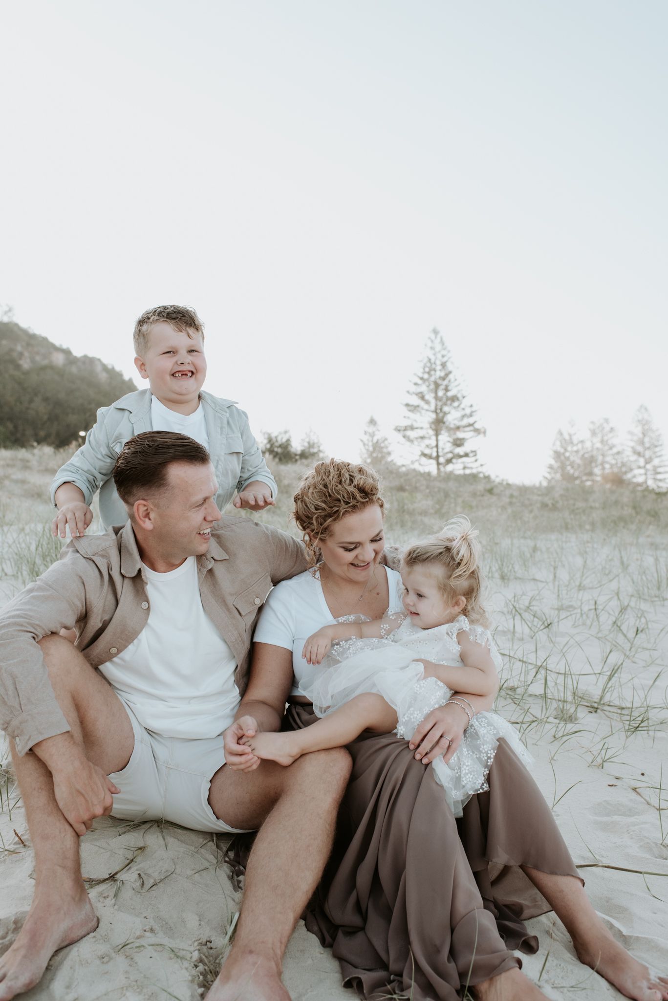 A family sitting together and laughing on a sandy beach with trees in the background at sunset.