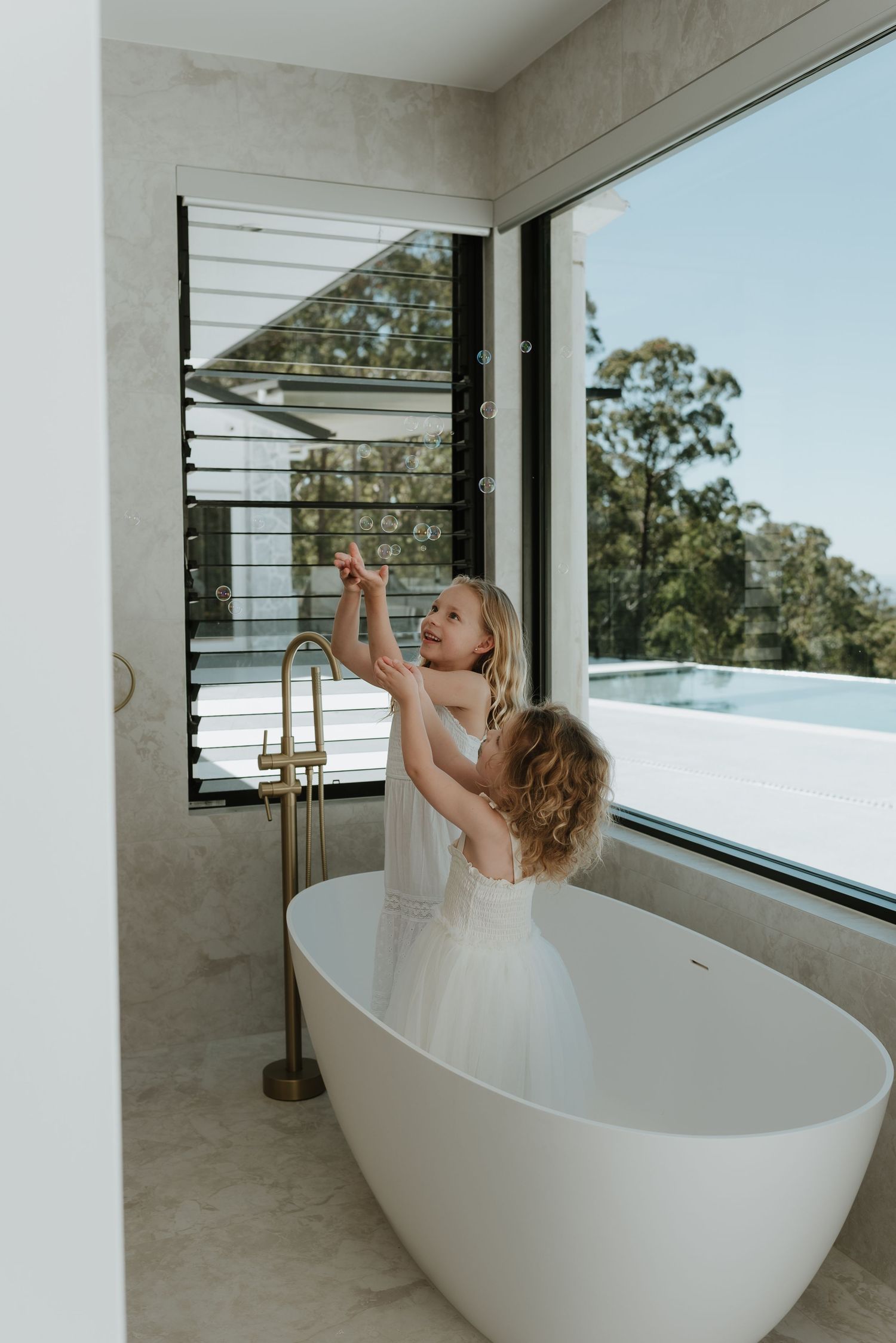 Two young girls in white dresses playing with bubbles beside a freestanding bathtub in a modern bathroom.