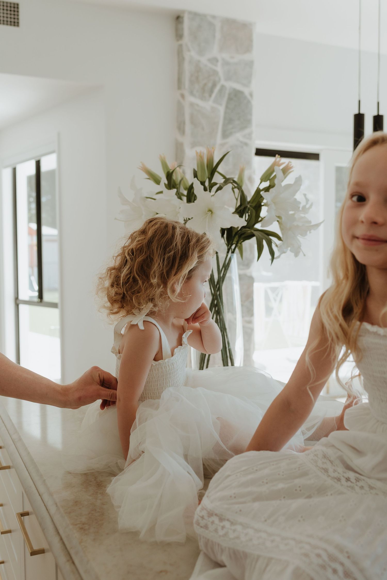 Two young girls in white flower girl dresses stand near a marble counter with white lilies in a bright, elegant room.