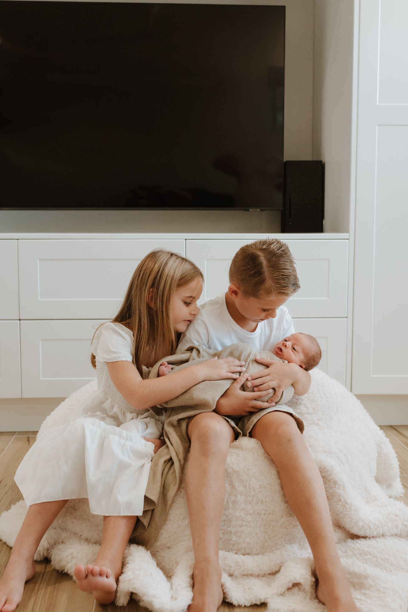 Two children sitting on a plush white rug while tenderly holding a newborn baby.