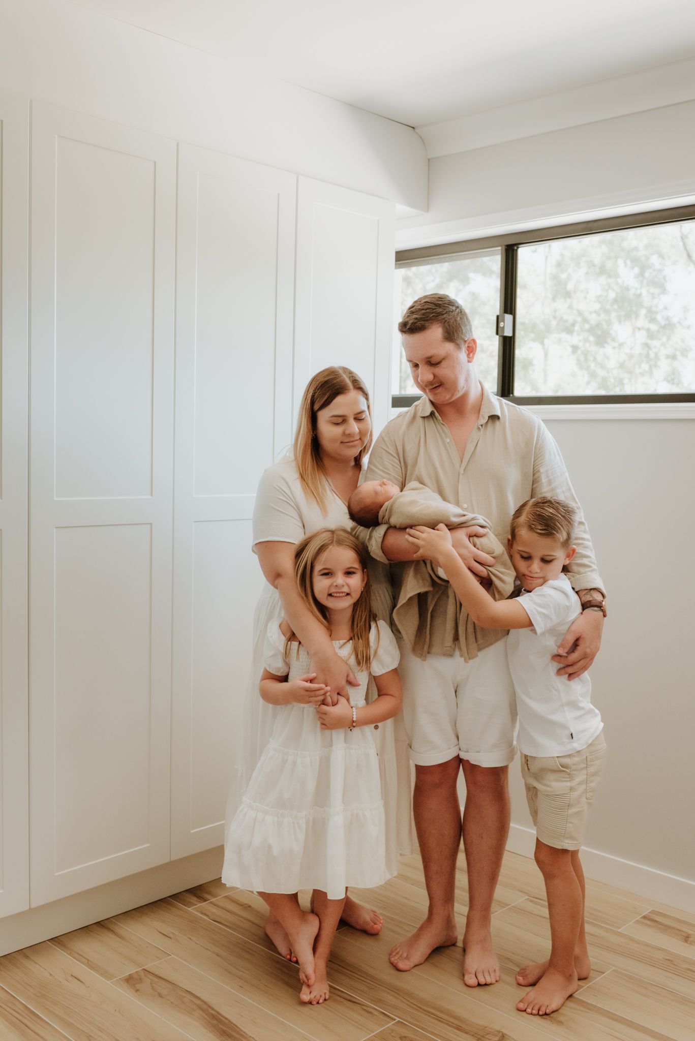 Family dressed in neutral tones standing together barefoot in bright minimalist room with wooden floors.