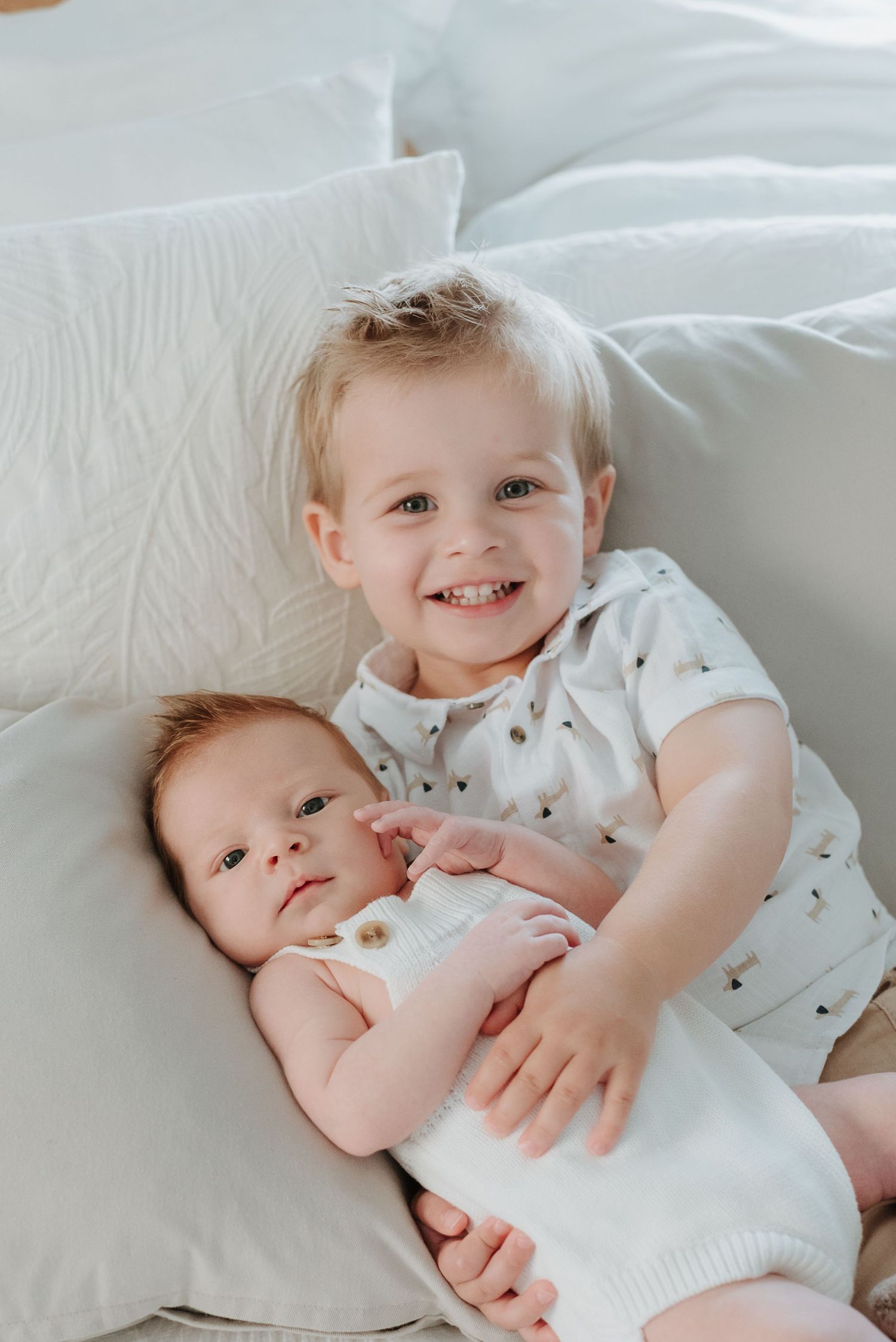 Young siblings wearing white outfits lie together on white bedding while sharing tender embraces and gentle moments.