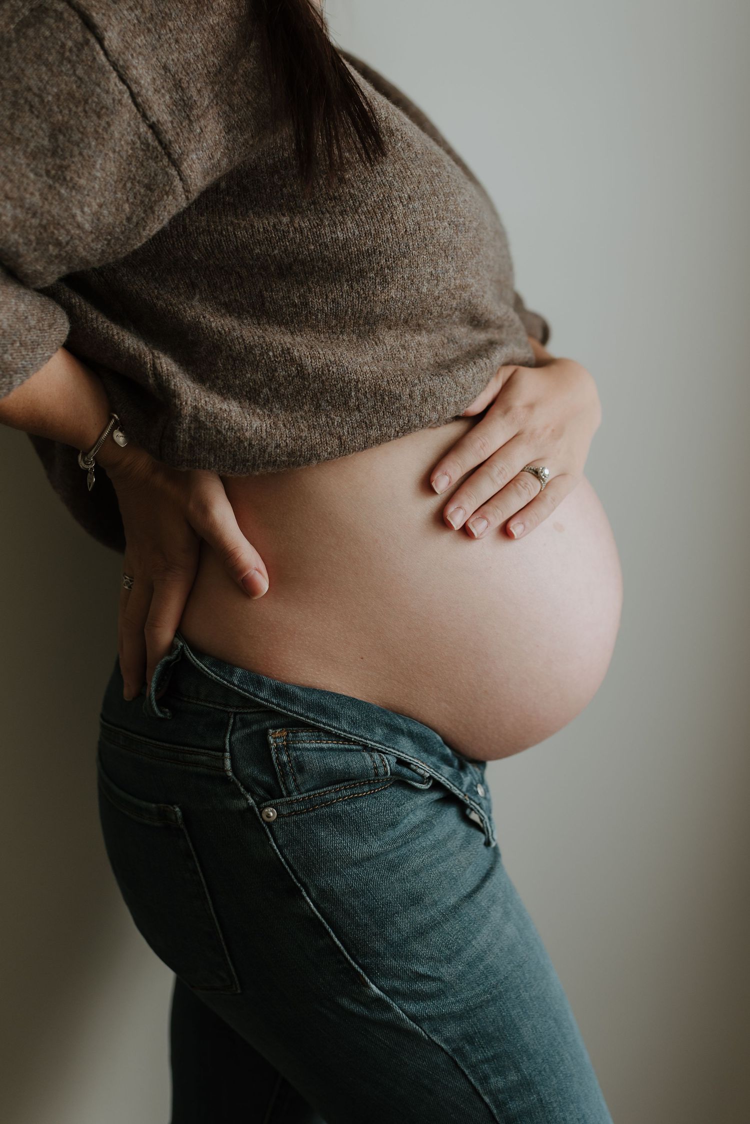 Pregnant woman in jeans and brown sweater cradling her bare baby bump with both hands against a neutral background.