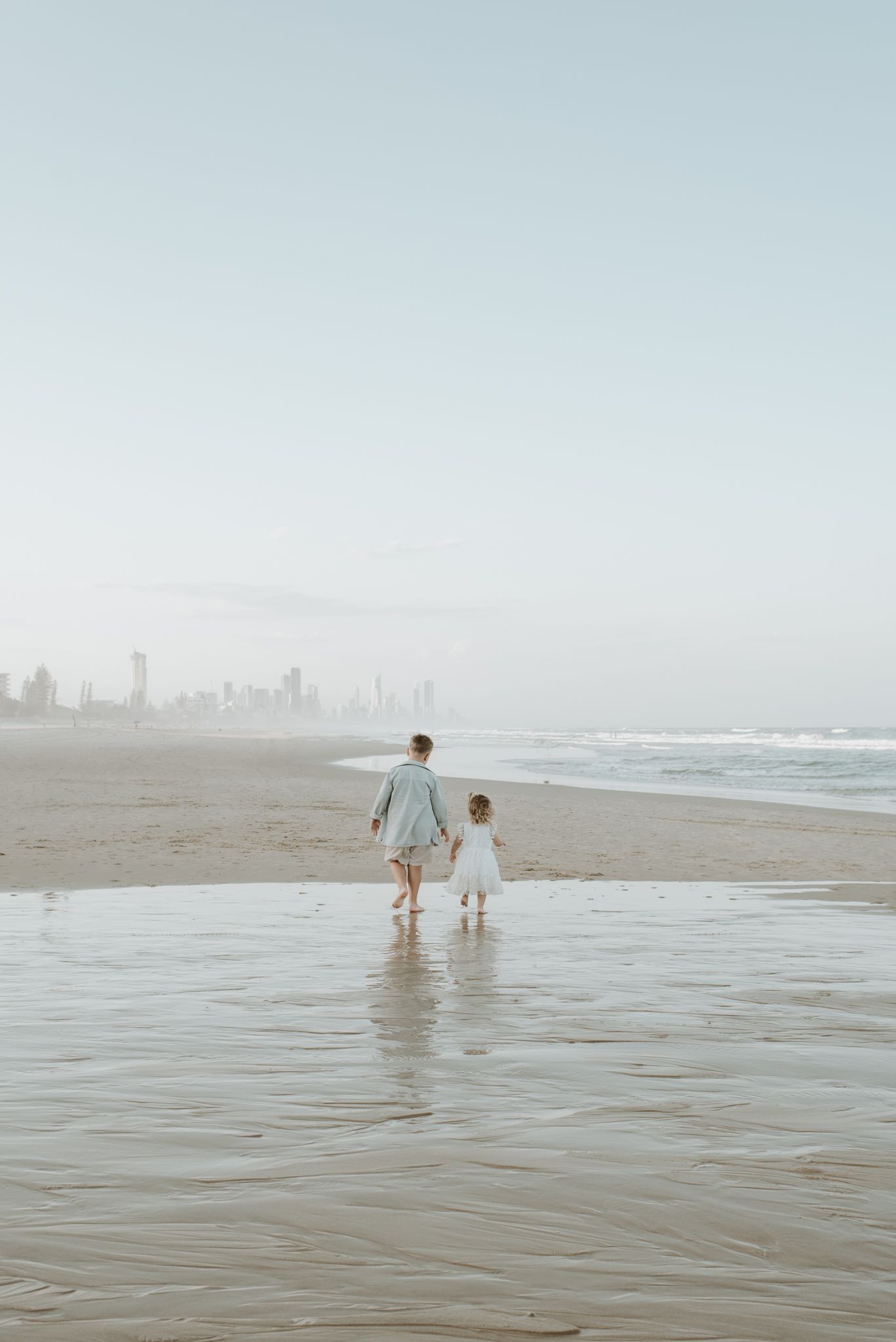 Two figures walking together on a misty beach with a city skyline visible in the distance.