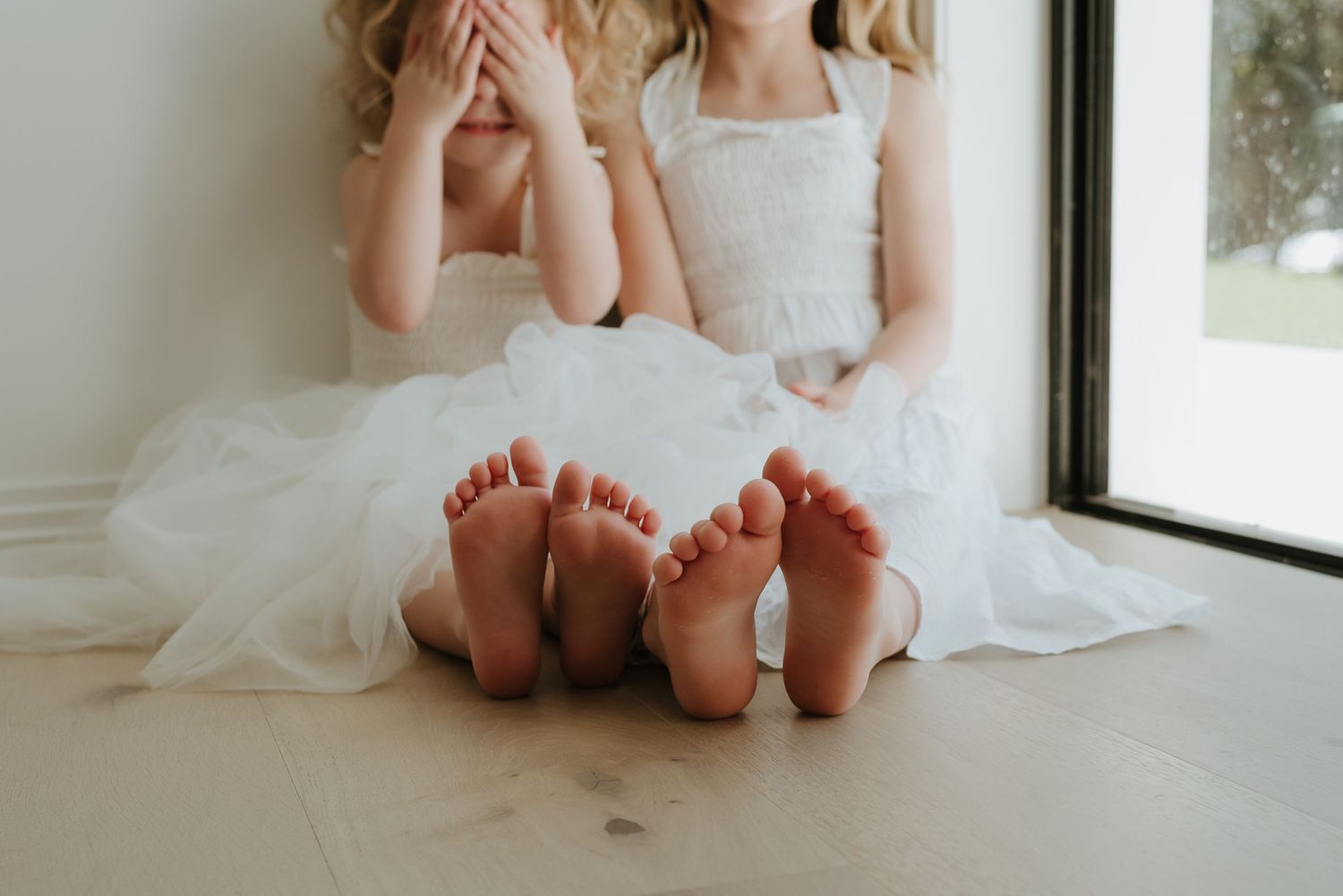 Two young girls in white tulle dresses sitting on a hardwood floor, showing their bare feet with painted toenails.