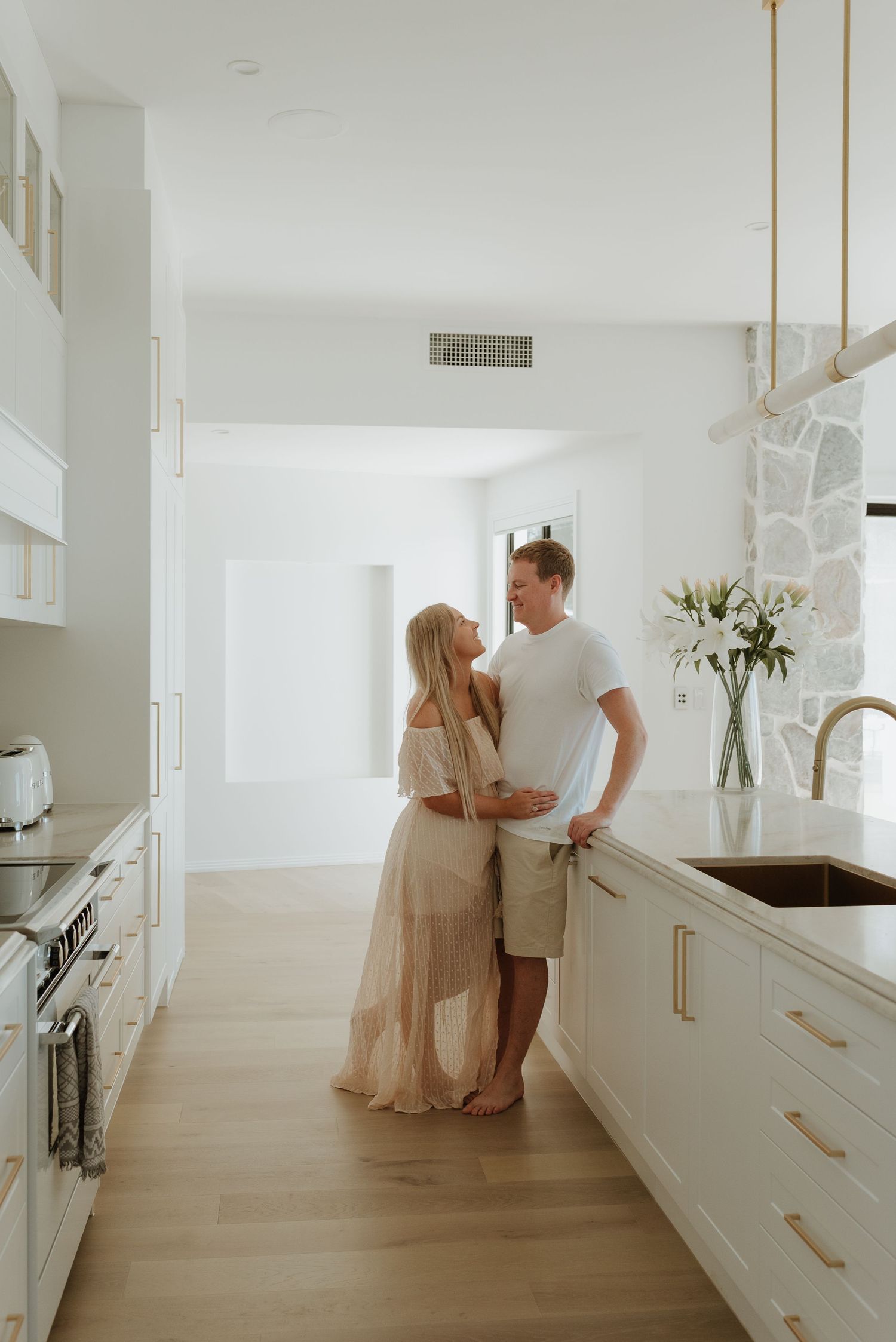 A couple embraces in a bright white modern kitchen with gold accents, hardwood floors, and fresh white flowers.