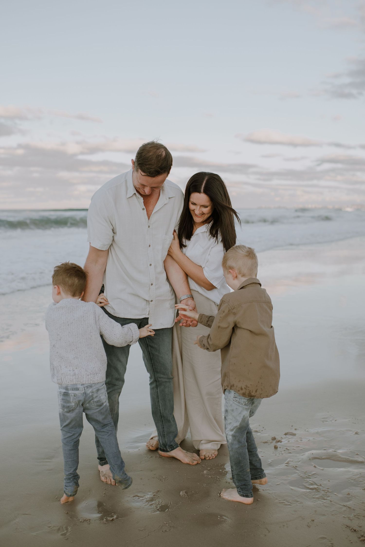A family holding hands while walking together on a sandy beach at sunset, wearing casual beige and white clothing.