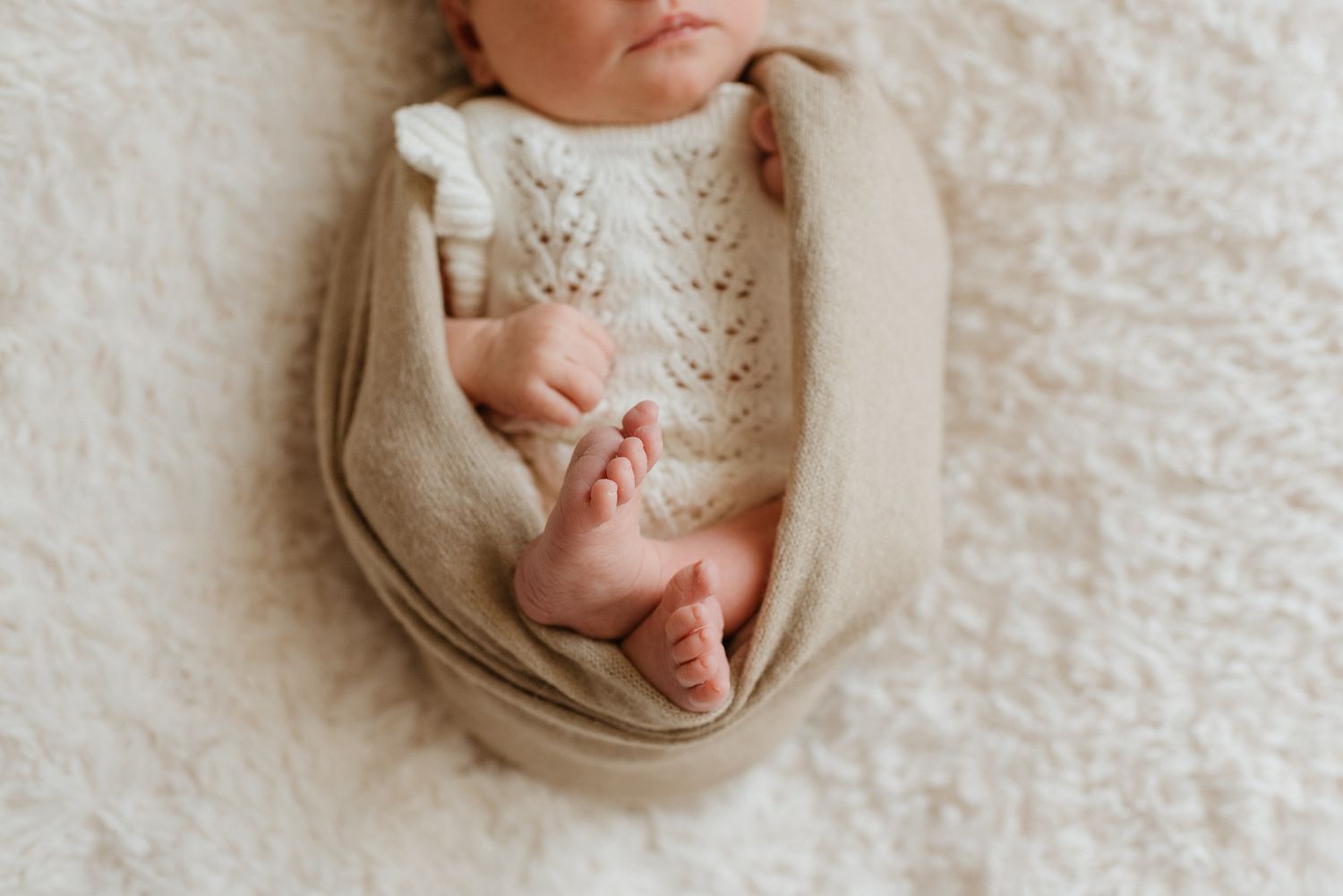 A sleeping newborn baby swaddled in beige fabric lying on a textured white blanket.