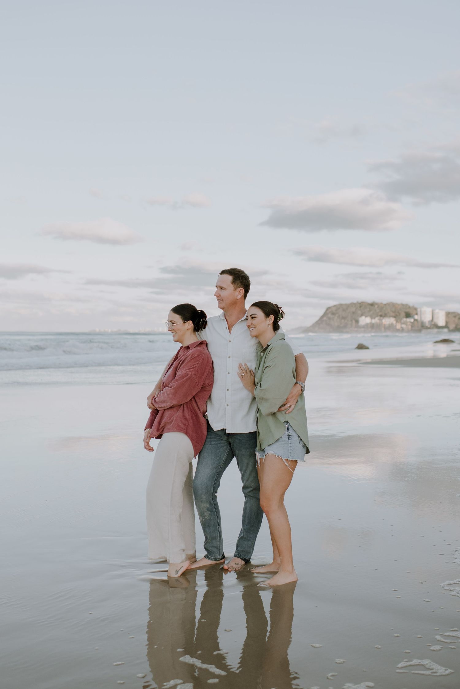 Three people stand together on a cloudy beach with mountains and buildings visible in the distance.
