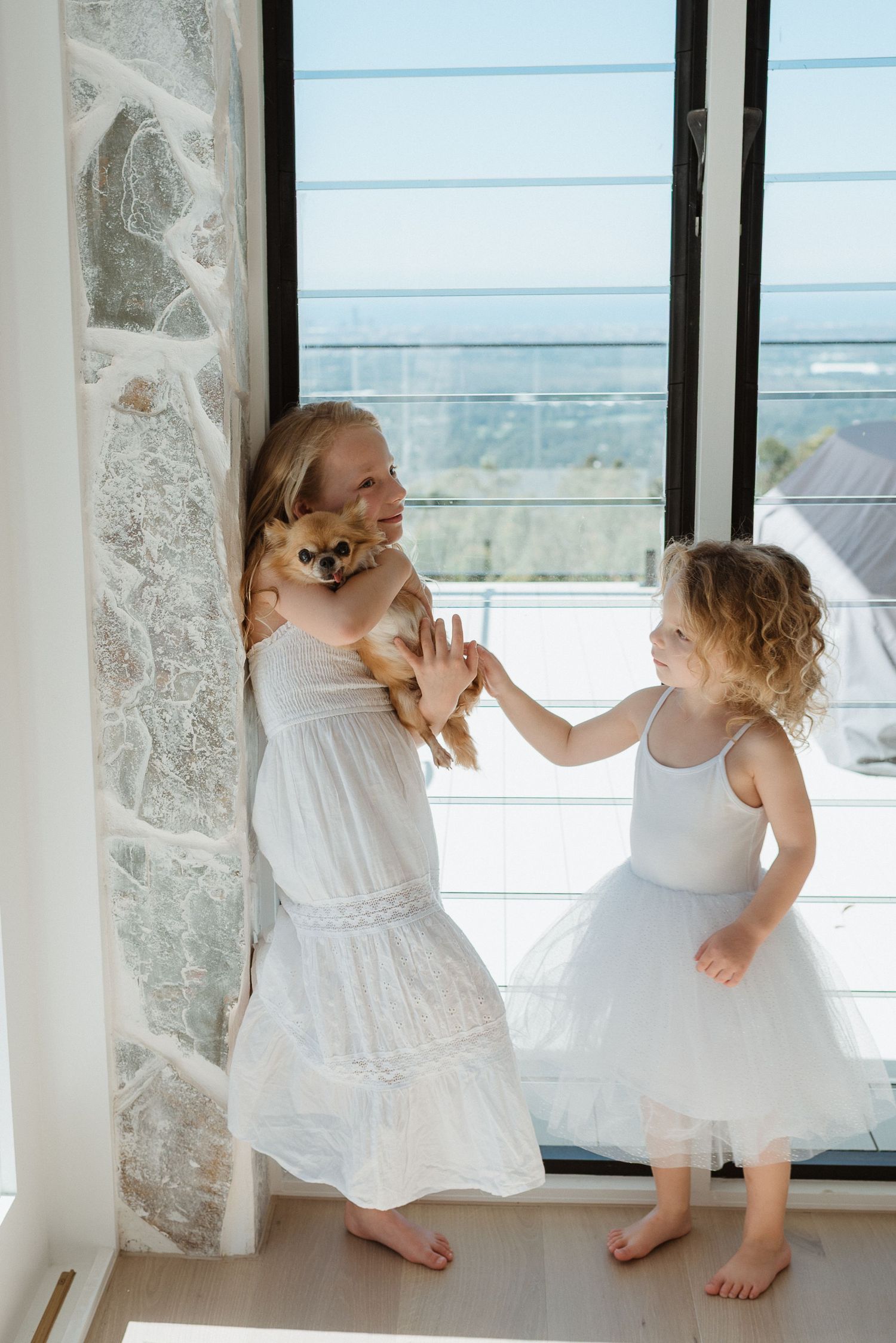 Two young girls in white dresses interact with a small brown dog near a sunlit window.