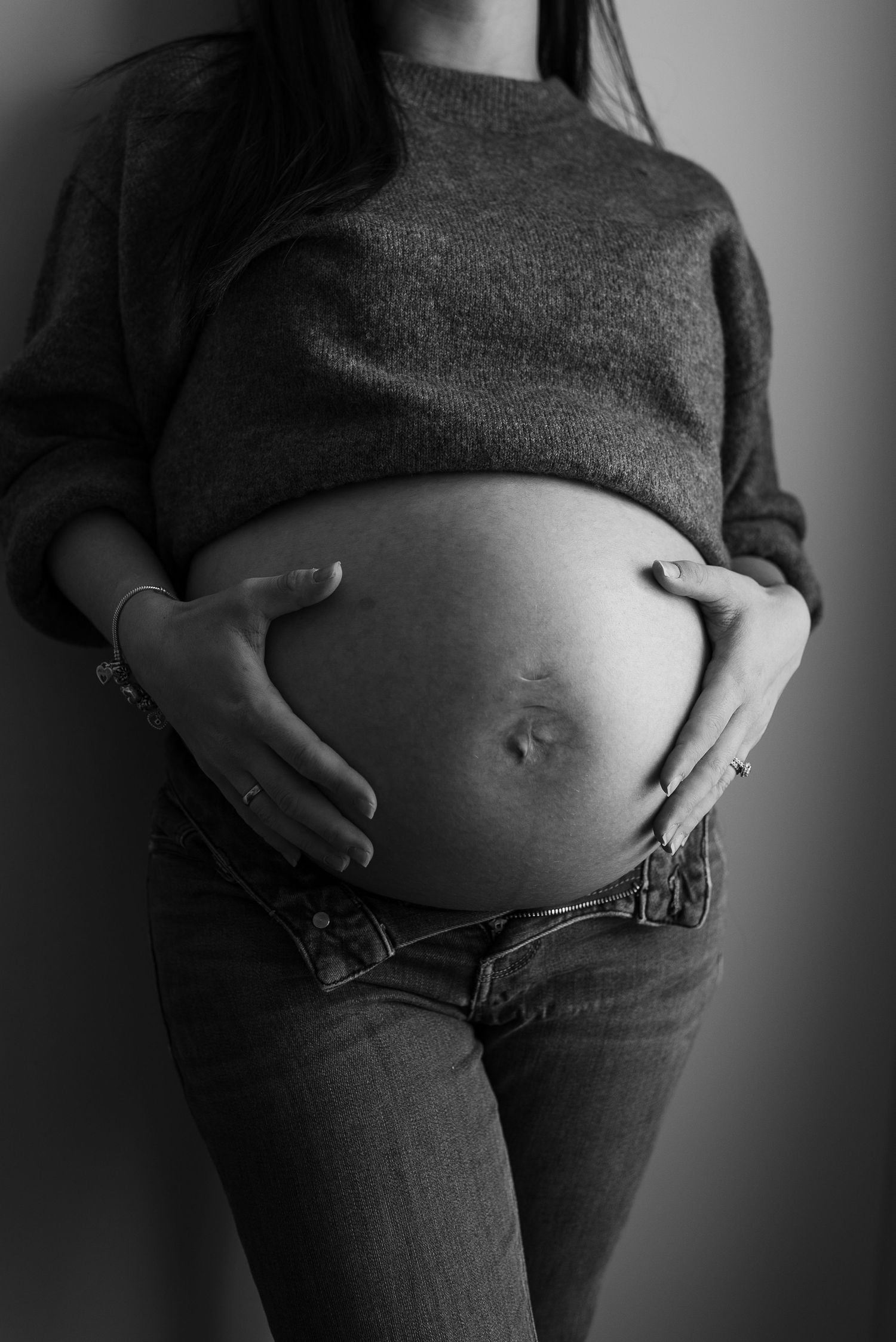 Black and white photo of a pregnant woman cradling her bare baby bump, wearing a gray sweater and jeans.