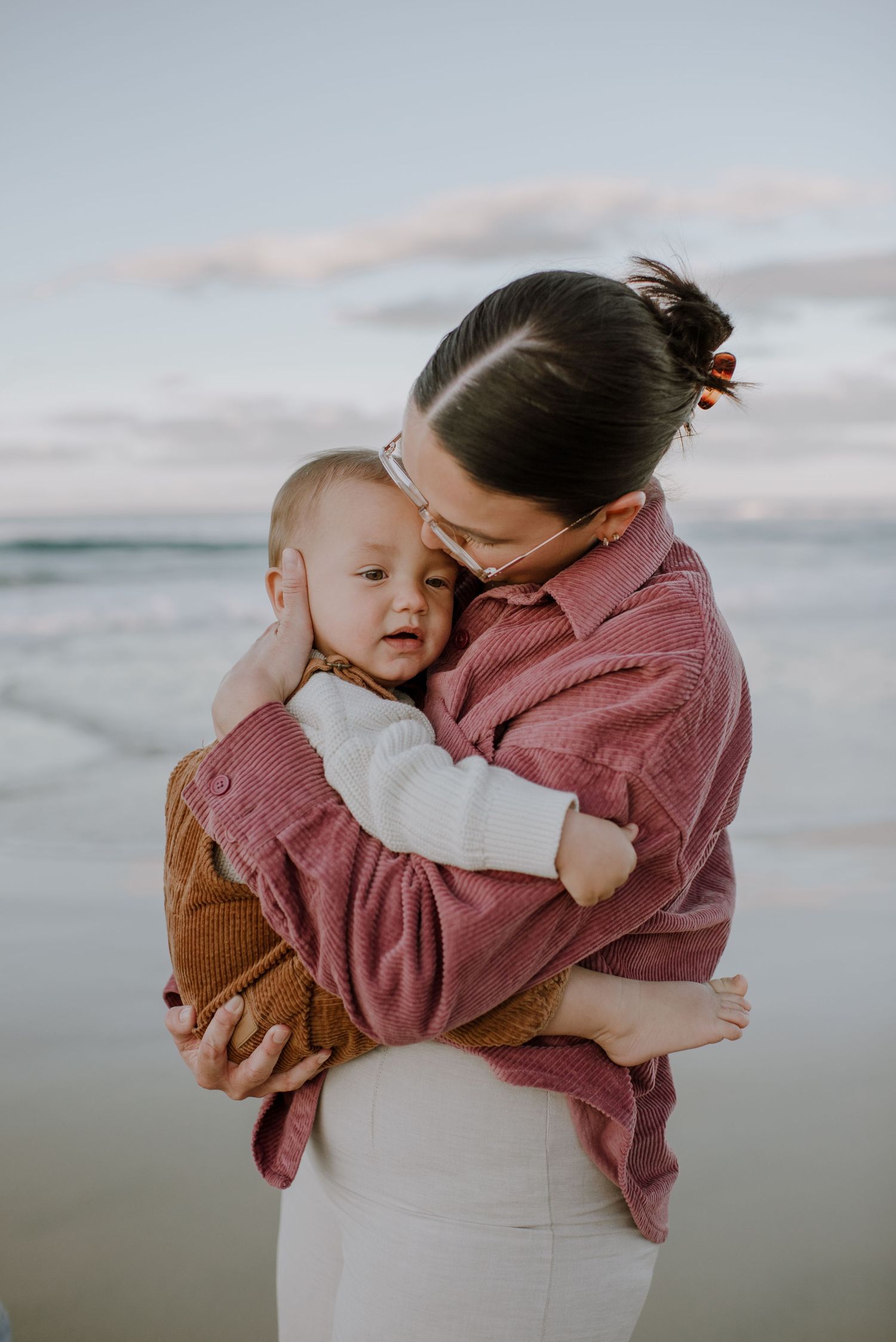 A tender moment shared between parent and baby at sunset on the beach, wearing cozy pink and white clothing.