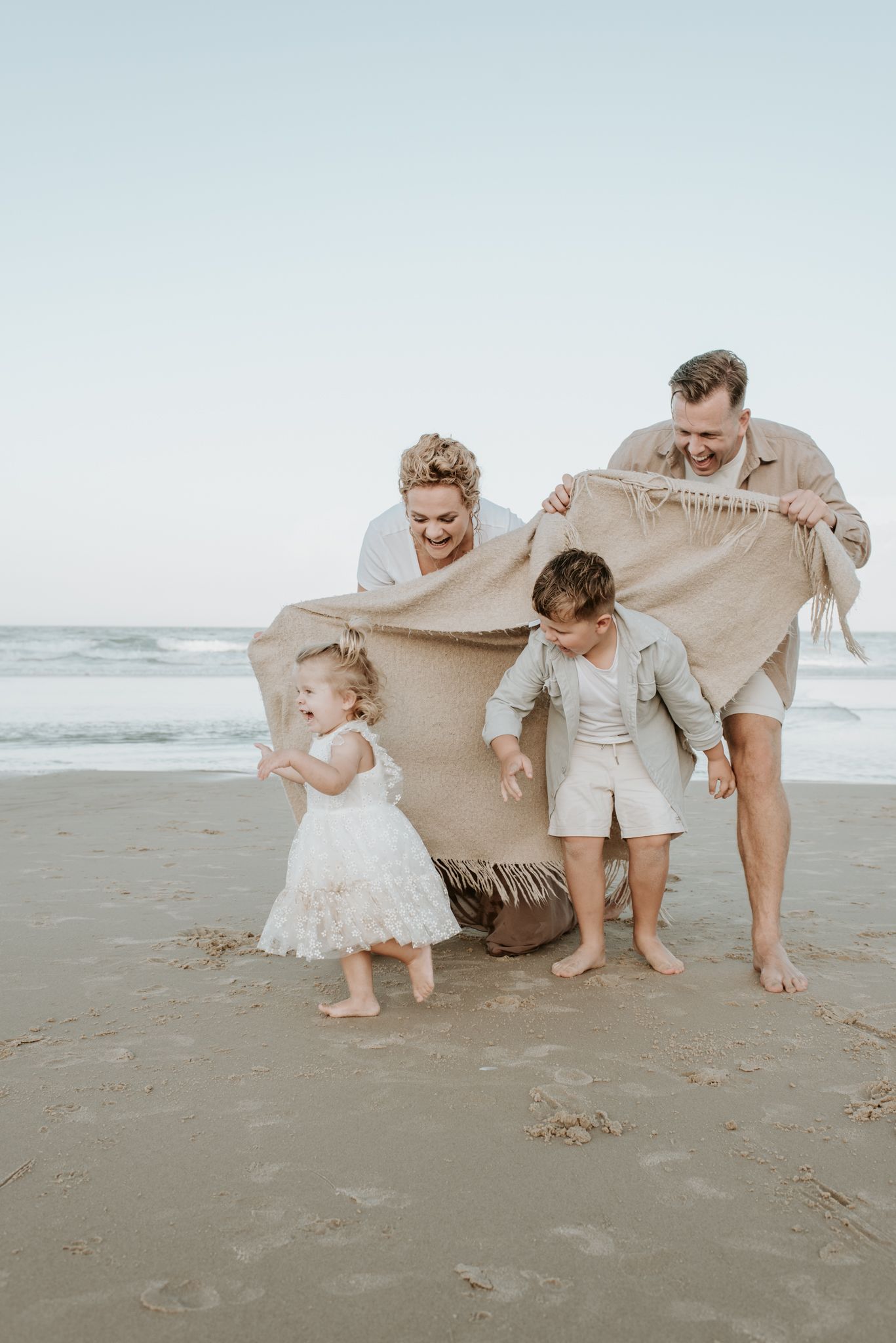 A playful family moment on the beach as they wrap themselves in a large beige blanket together.