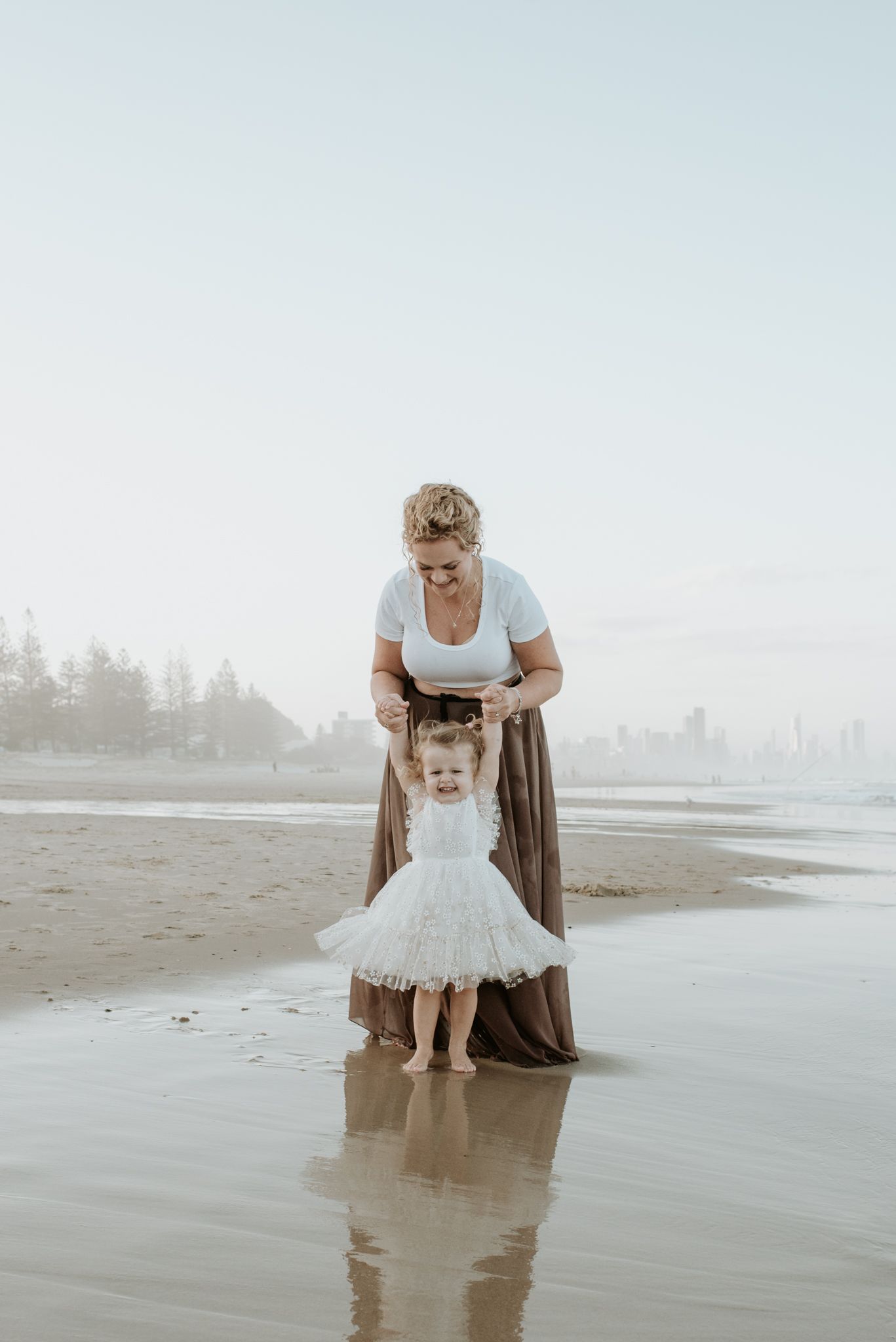 A tender moment captured on a misty beach as they stand together in the wet sand with their reflections visible below.