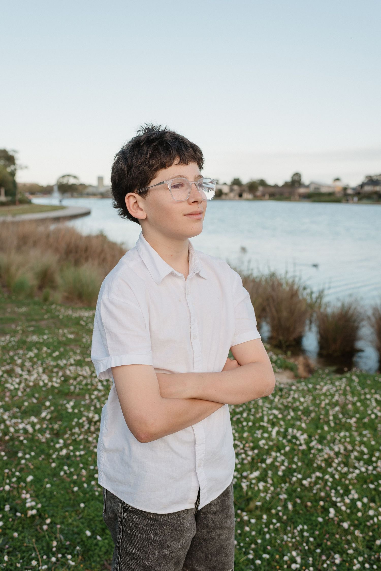 A teenage boy with glasses and crossed arms stands in a flower field near a lake, wearing a white button-up shirt.