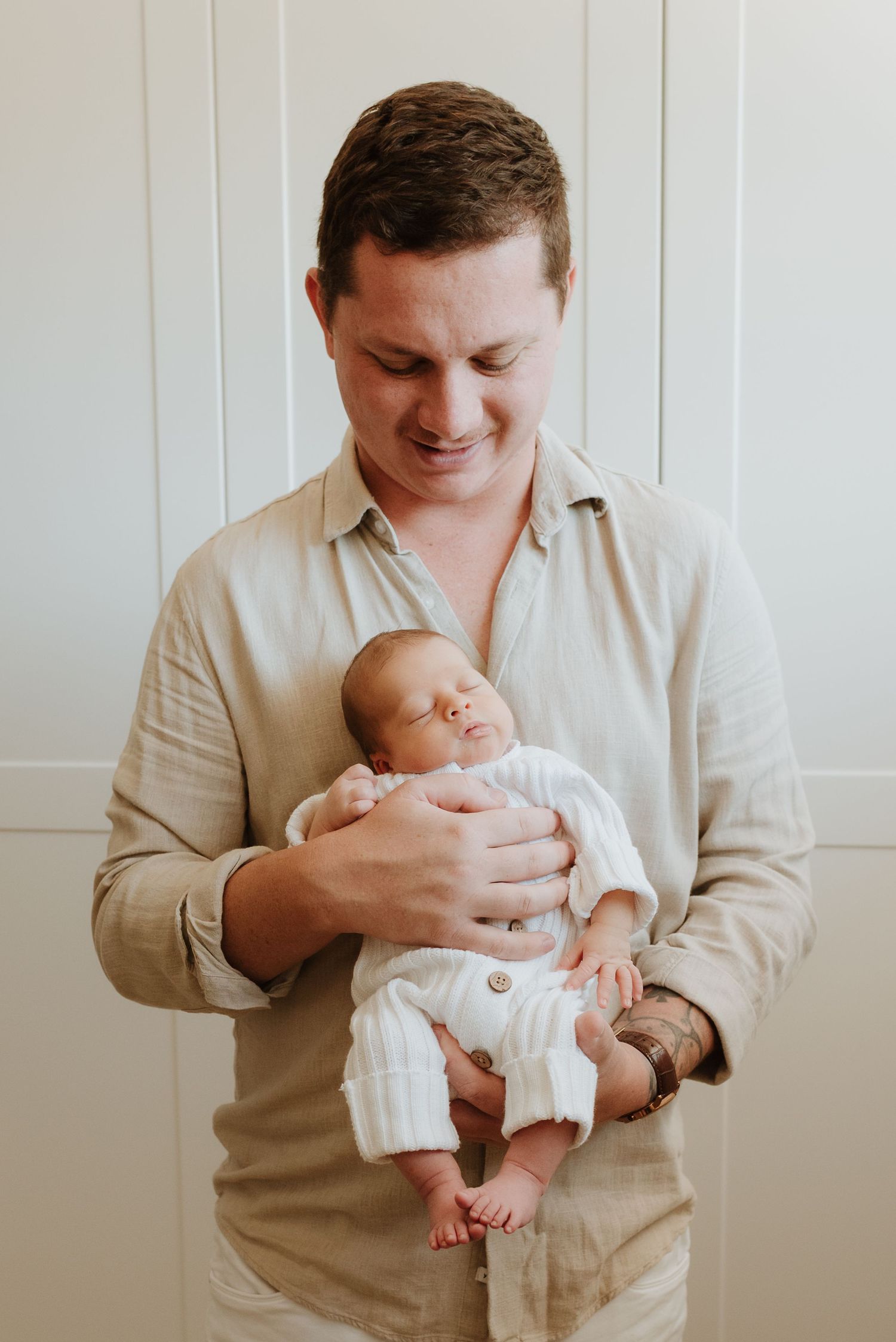 A parent in a beige shirt cradles a newborn baby in a series of tender moments against a white wall background.