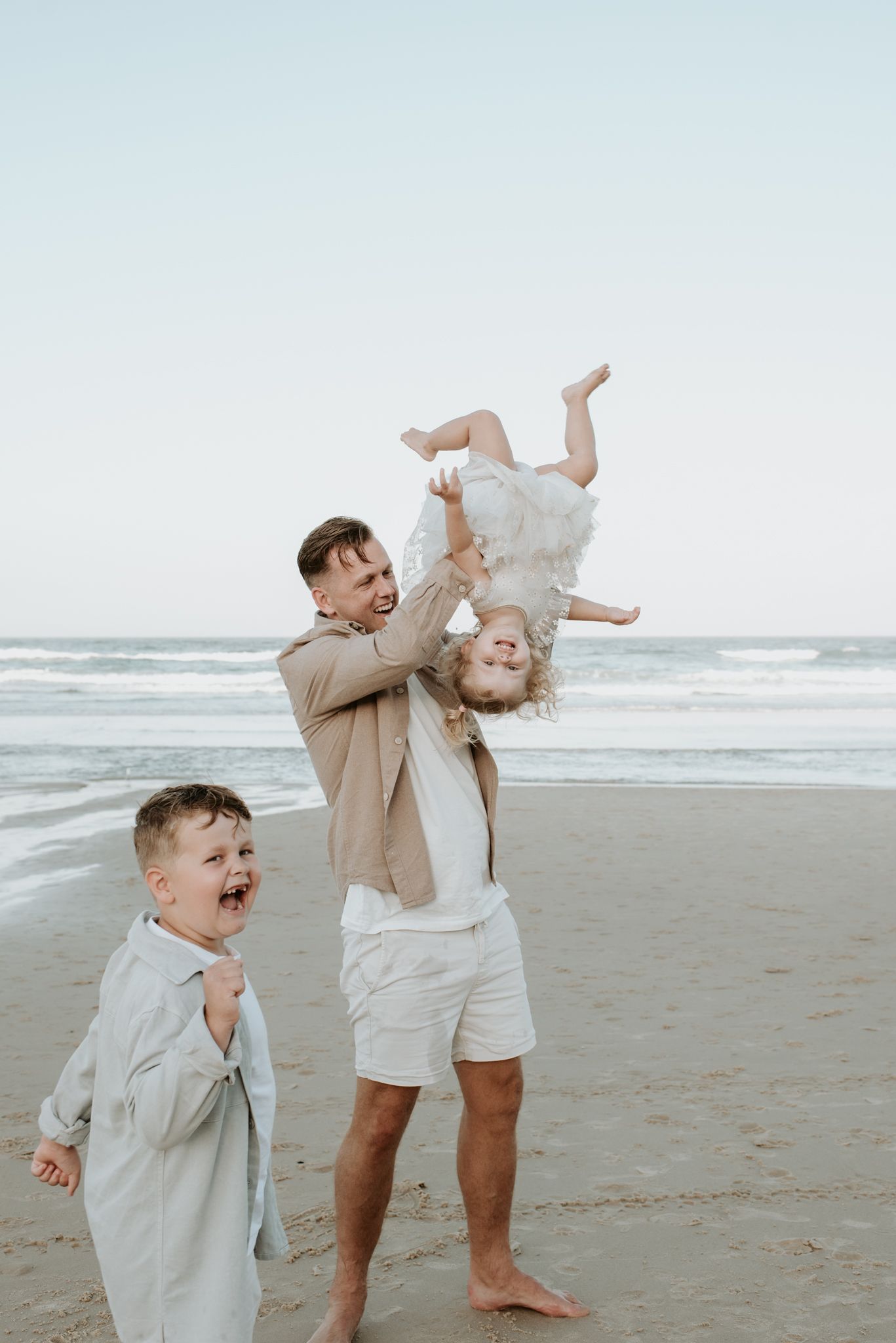 Playful family moment captured on a cloudy beach with joyful laughter and a child being lifted in the air.