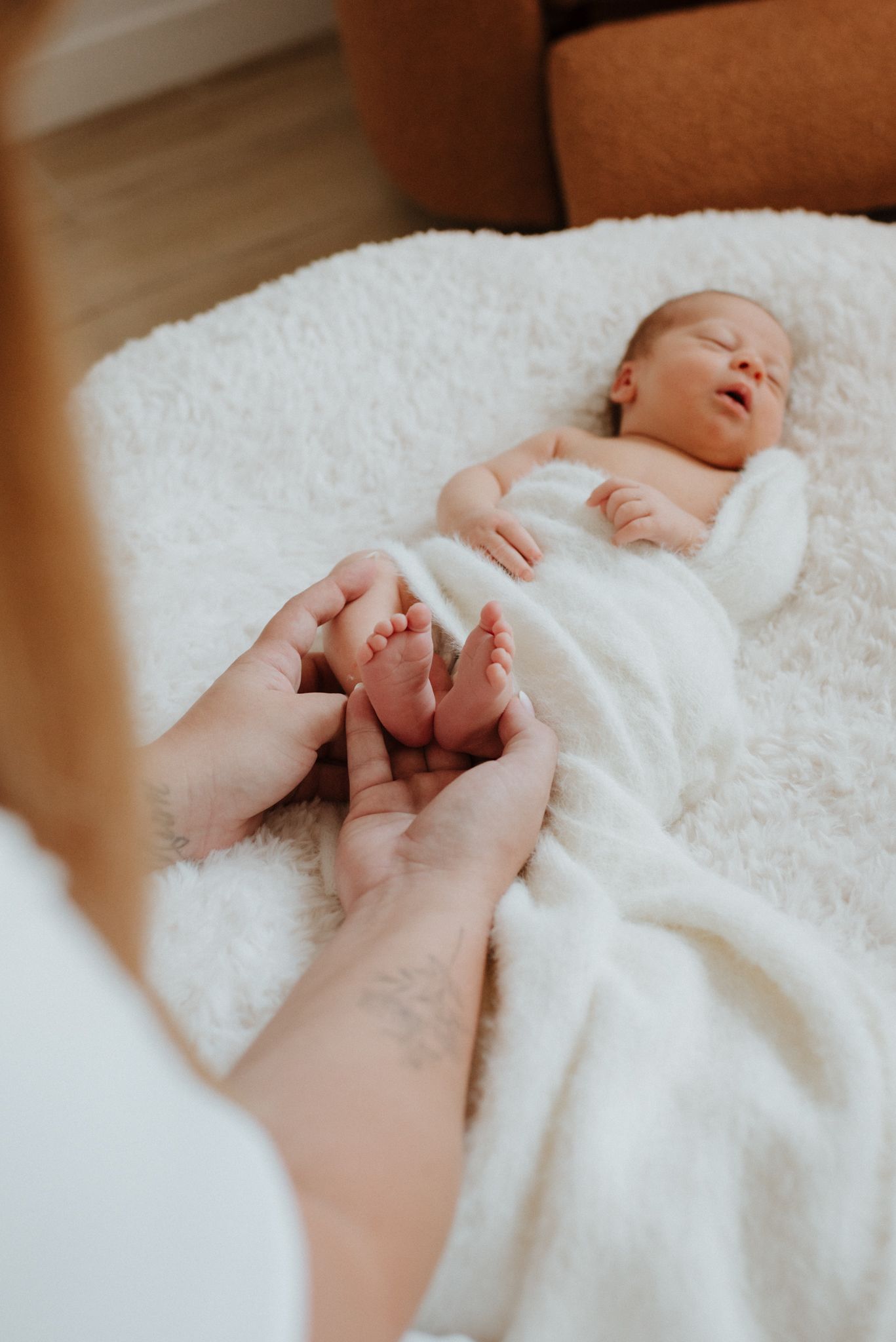 Gentle hands touching tiny feet of newborn baby wrapped in soft white blanket resting on plush surface.
